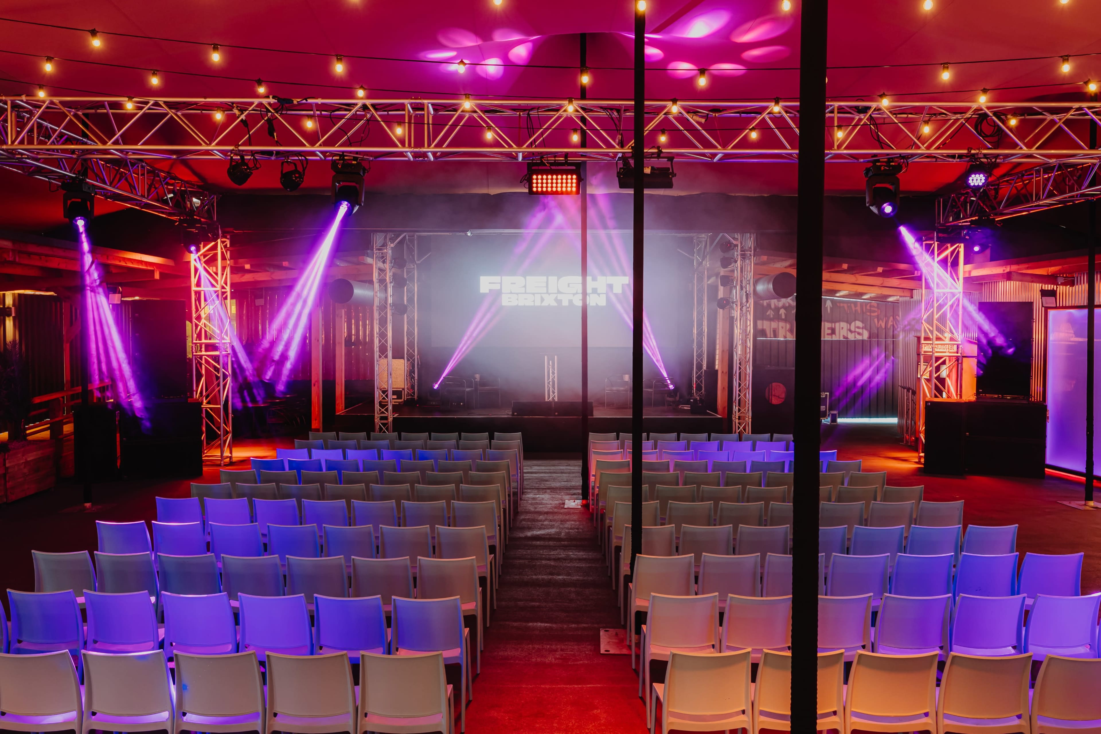 The image shows an event space set up for a performance, featuring rows of empty chairs facing a stage illuminated by colorful lights and a large screen.
