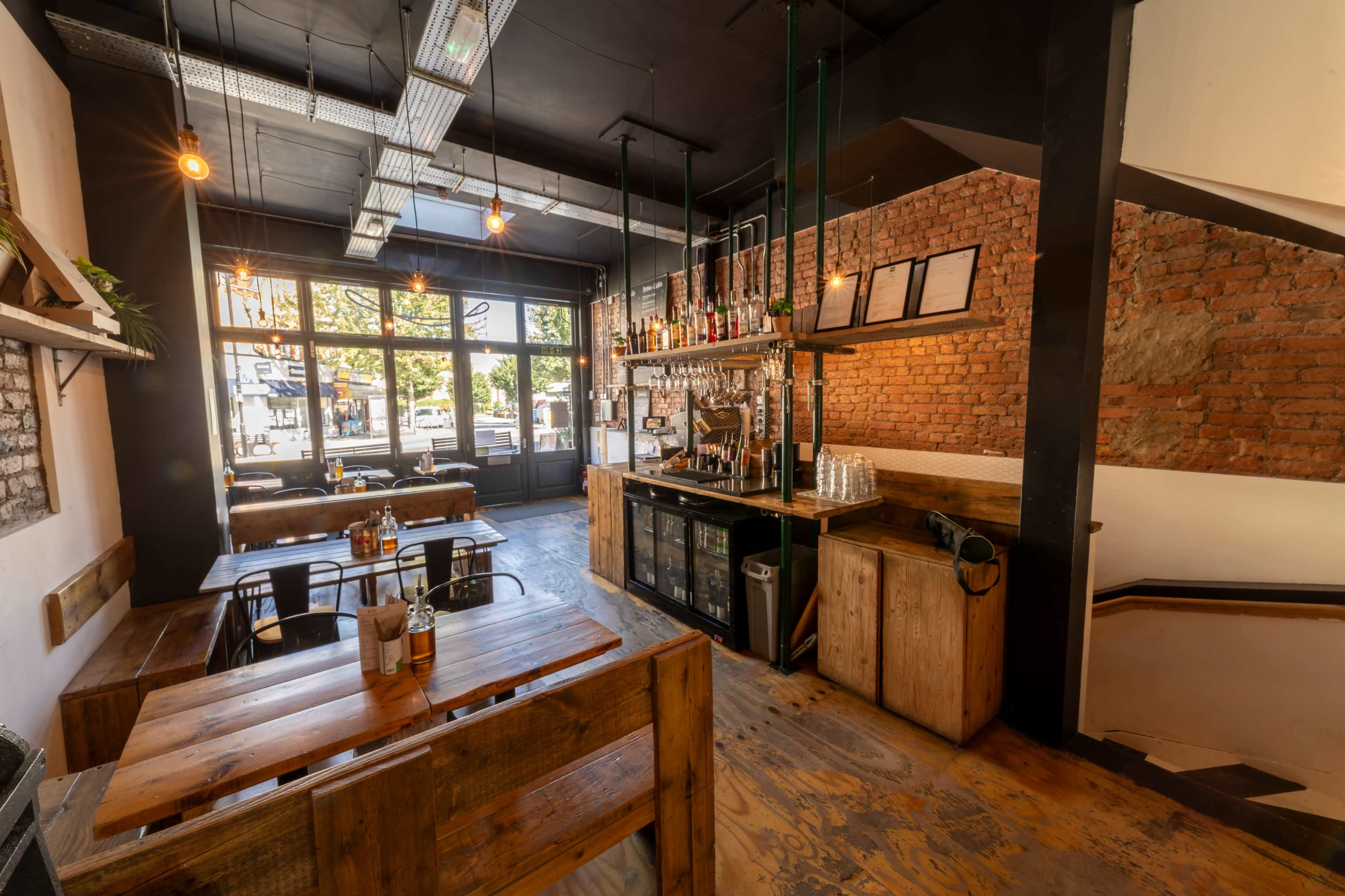 The image shows a modern cafe with exposed brick walls, wooden furniture, and a bar area featuring various bottles and bar equipment.