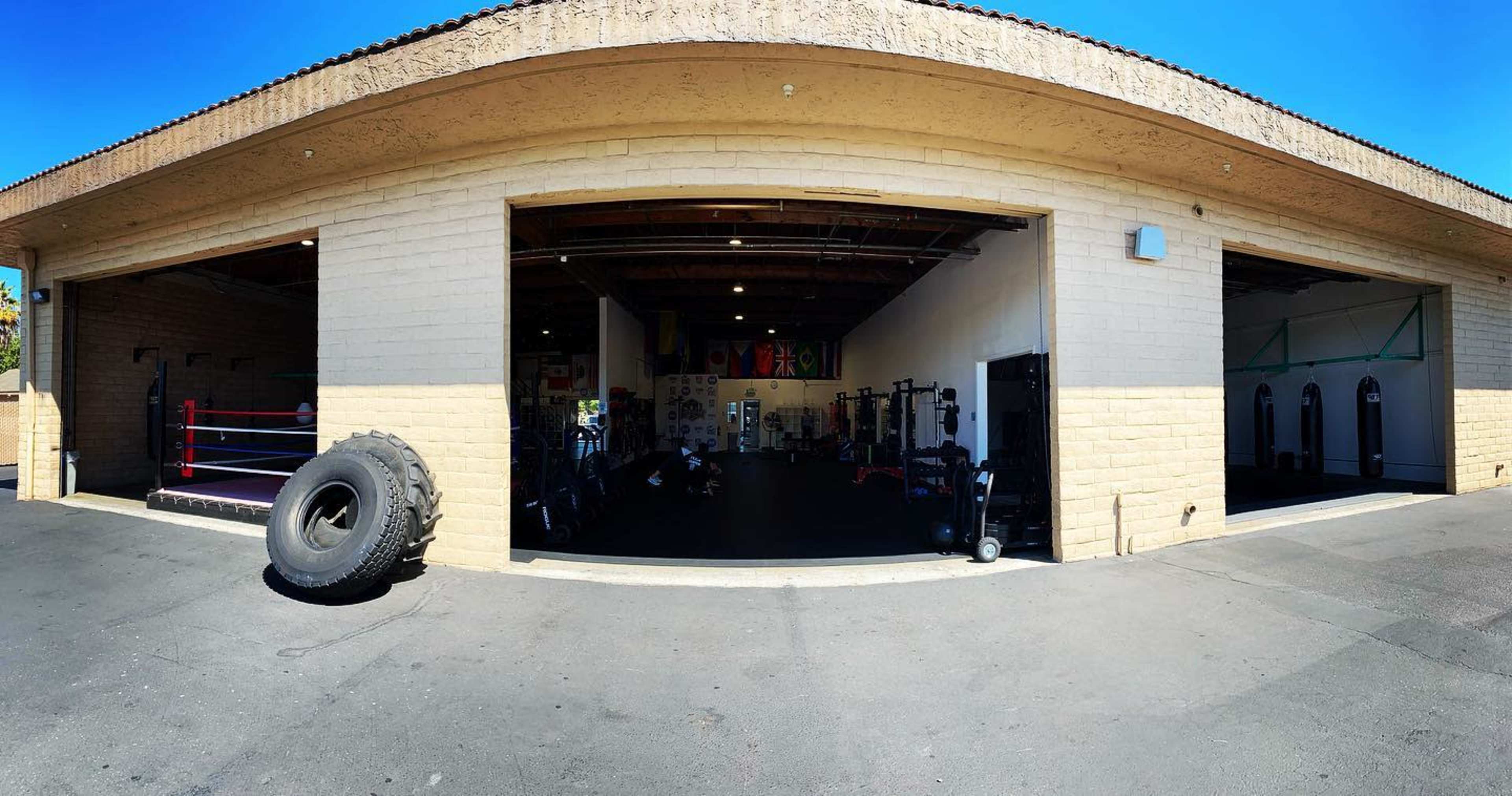 A spacious gym with large open doors, featuring workout equipment, a boxing ring, and a large tire outside.