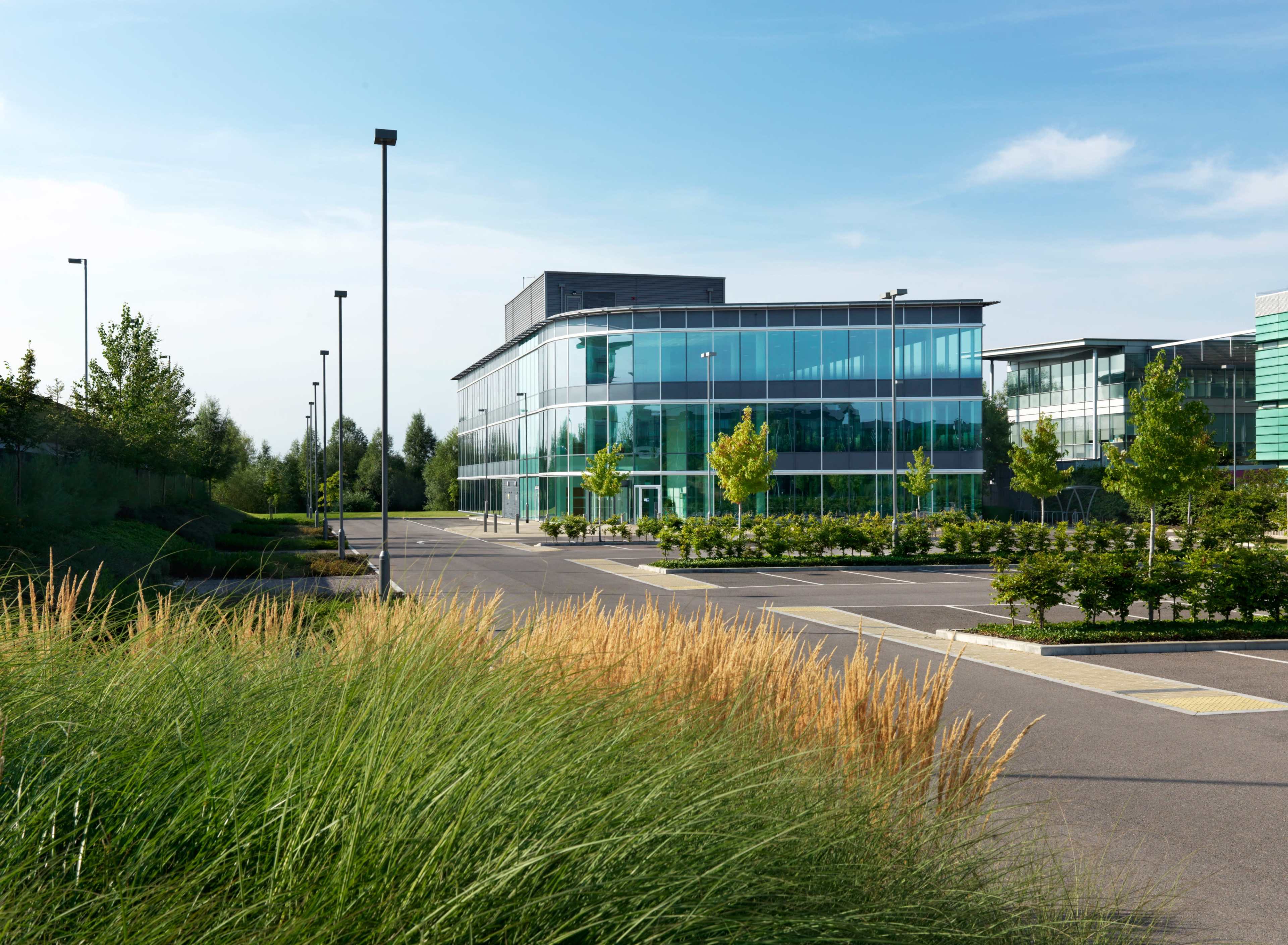 A modern glass-fronted office building is situated beside a landscaped pathway with tall grass and trees.
