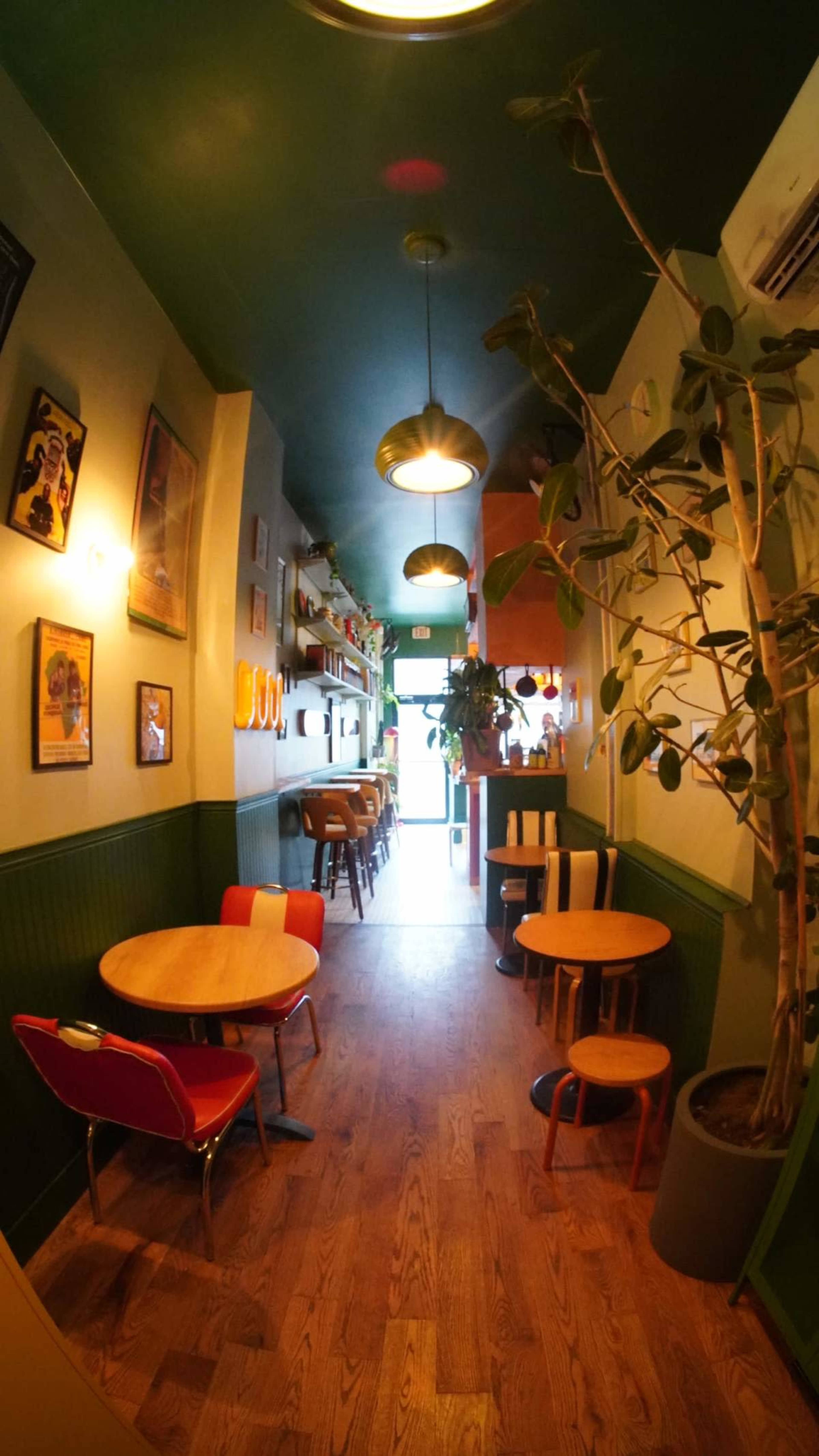 The image shows a narrow café hallway with wooden flooring, small round tables, and greenery, leading to a brightly lit entrance.