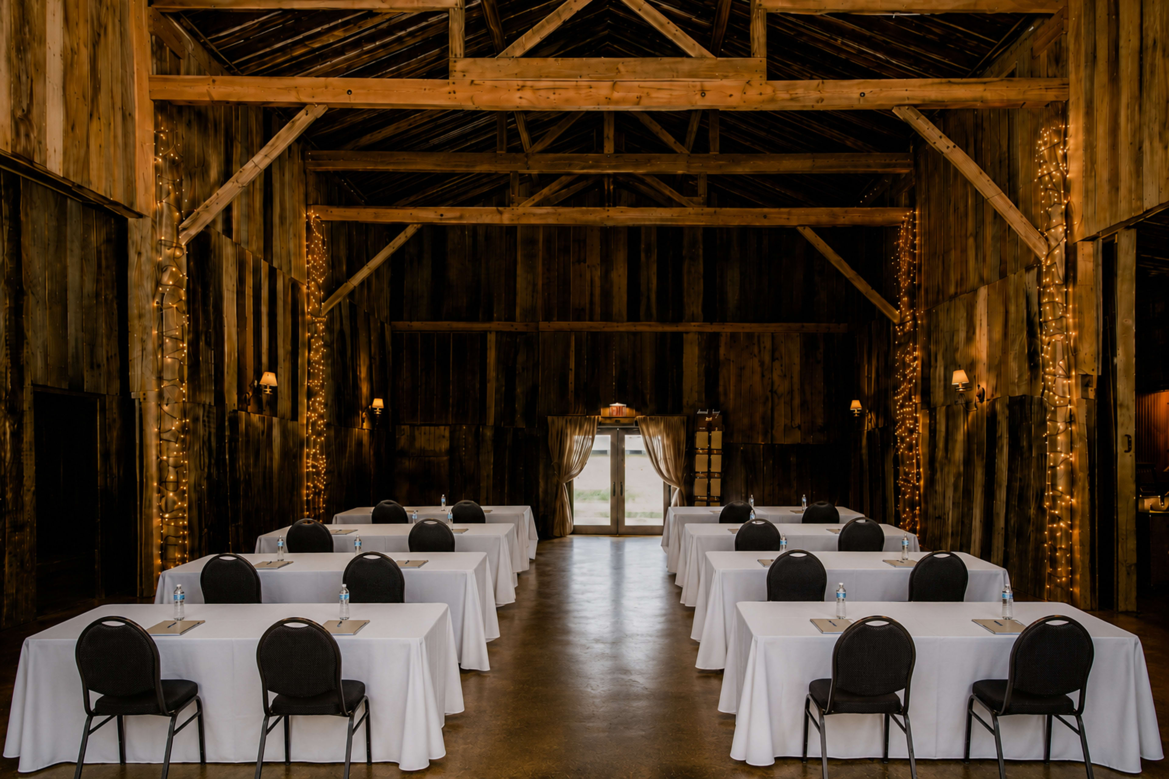 The image shows a rustic barn interior set up for an event, featuring rows of tables covered in white tablecloths and surrounded by black chairs, with string lights adorning the wooden walls.