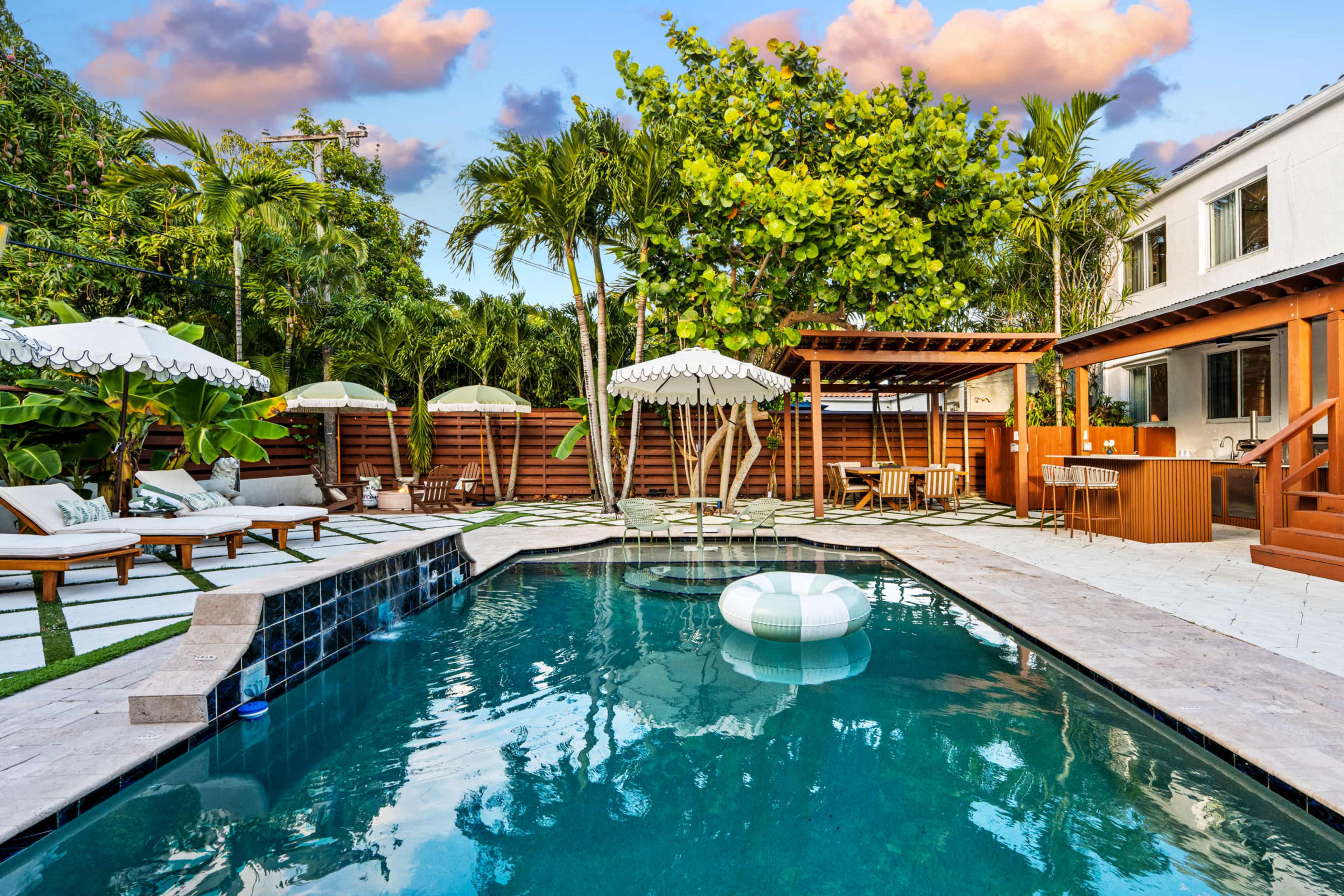 The image shows a well-designed backyard with a swimming pool, lounge chairs, umbrellas, and a wooden deck surrounded by tropical plants and trees.