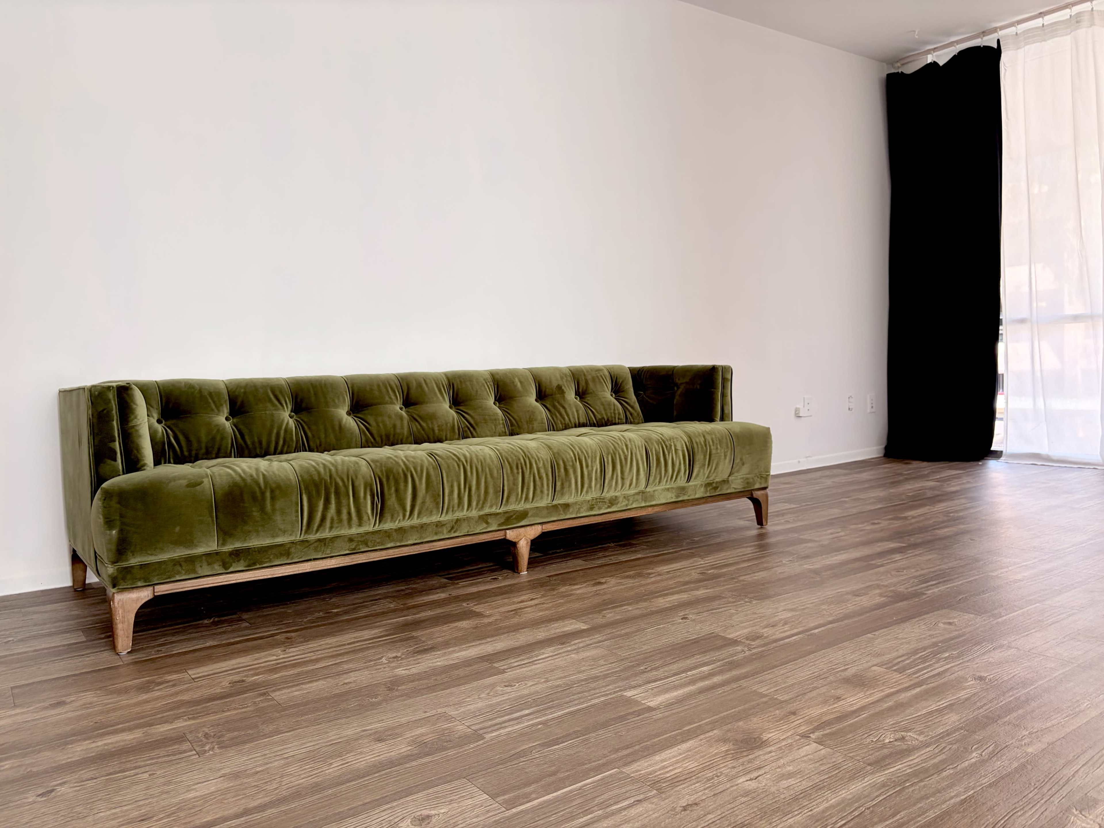A green velvet sofa with a tufted design is placed against a plain white wall in a room with wooden flooring and a large window covered by sheer curtains.