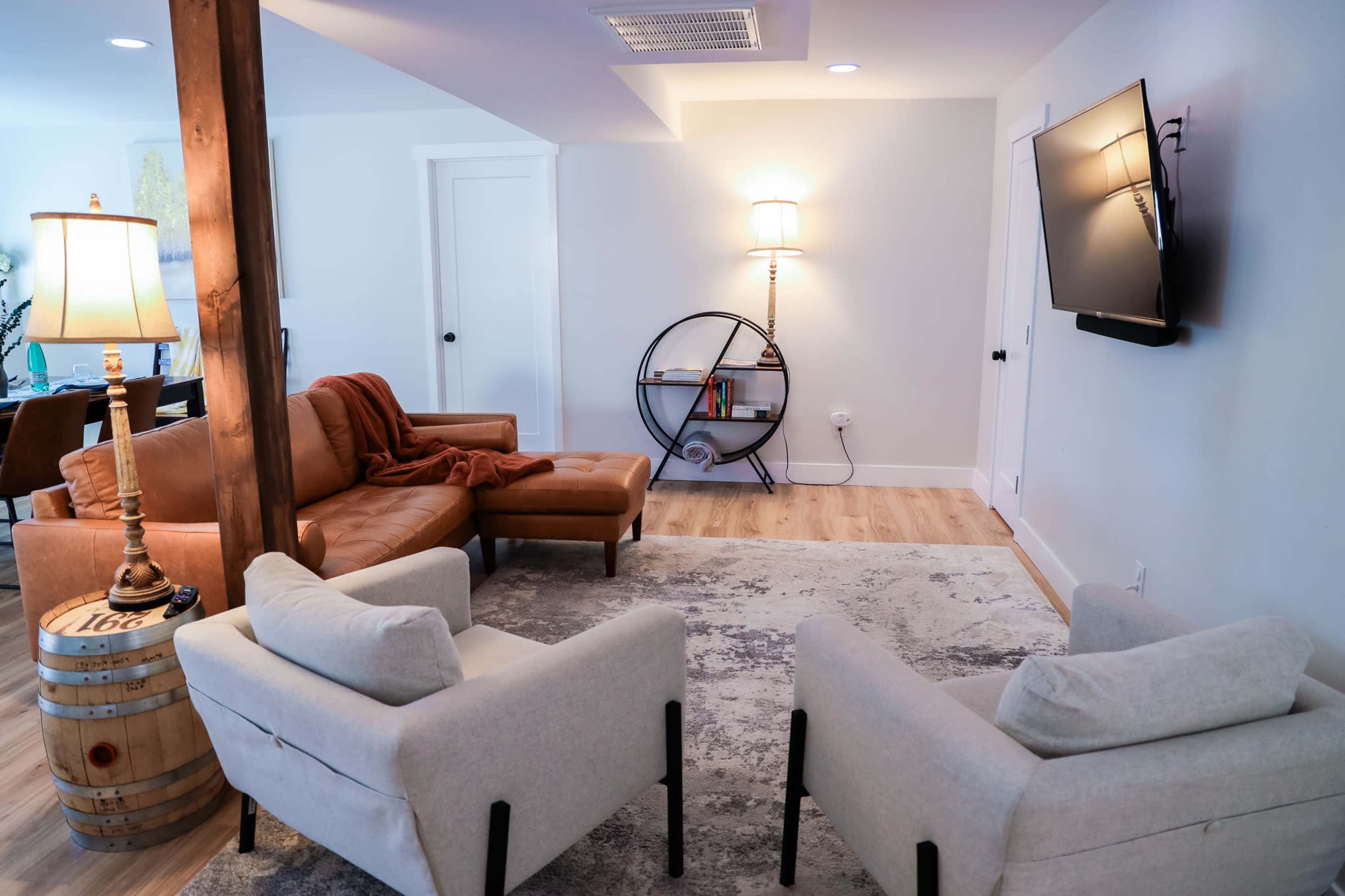 A cozy living room features a brown leather sofa, two light-colored armchairs, a wooden barrel, a round bookshelf, and a wall-mounted television.