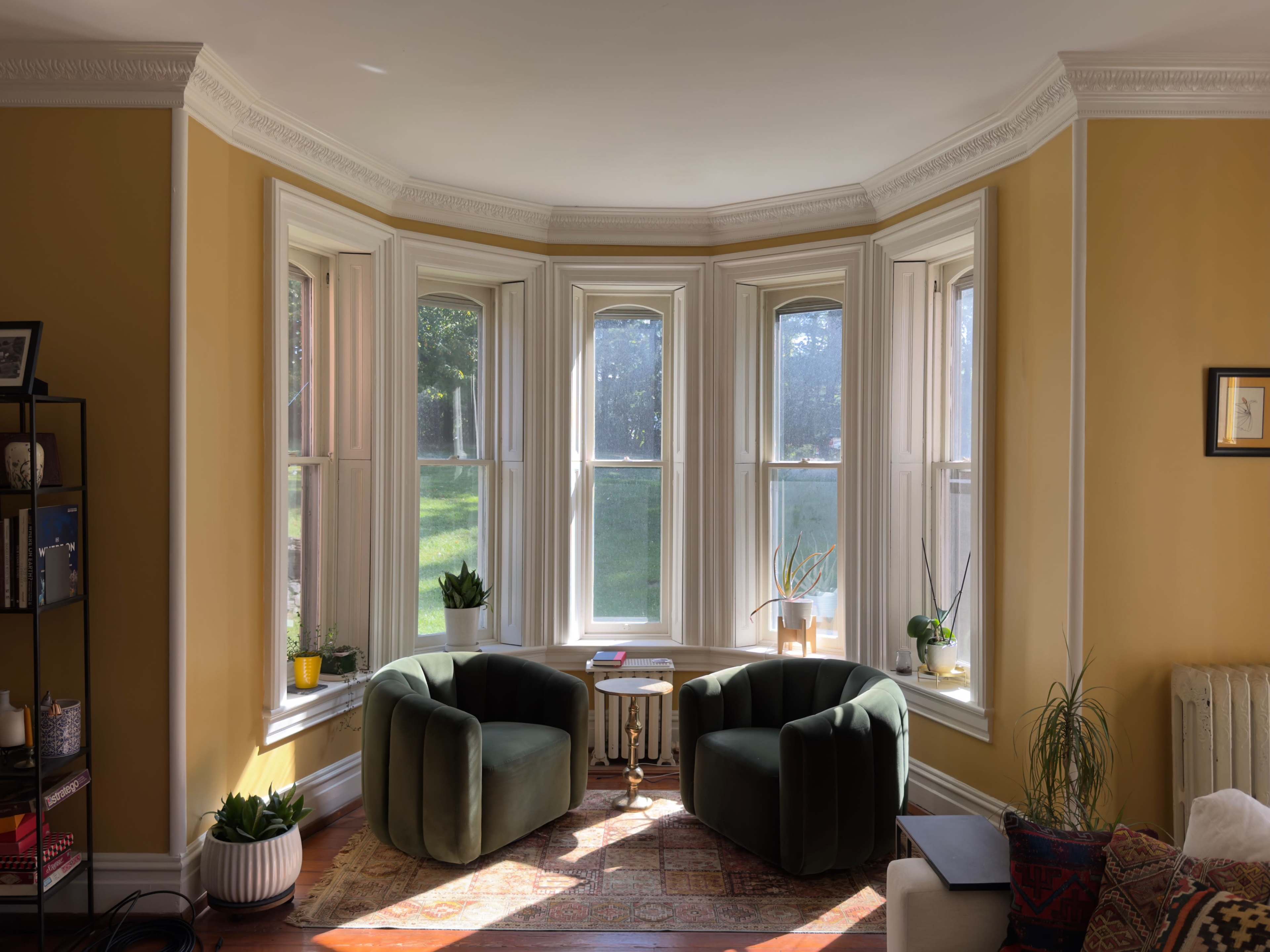 The image shows a cozy living room with two green chairs facing a bay window, surrounded by plants and natural light.