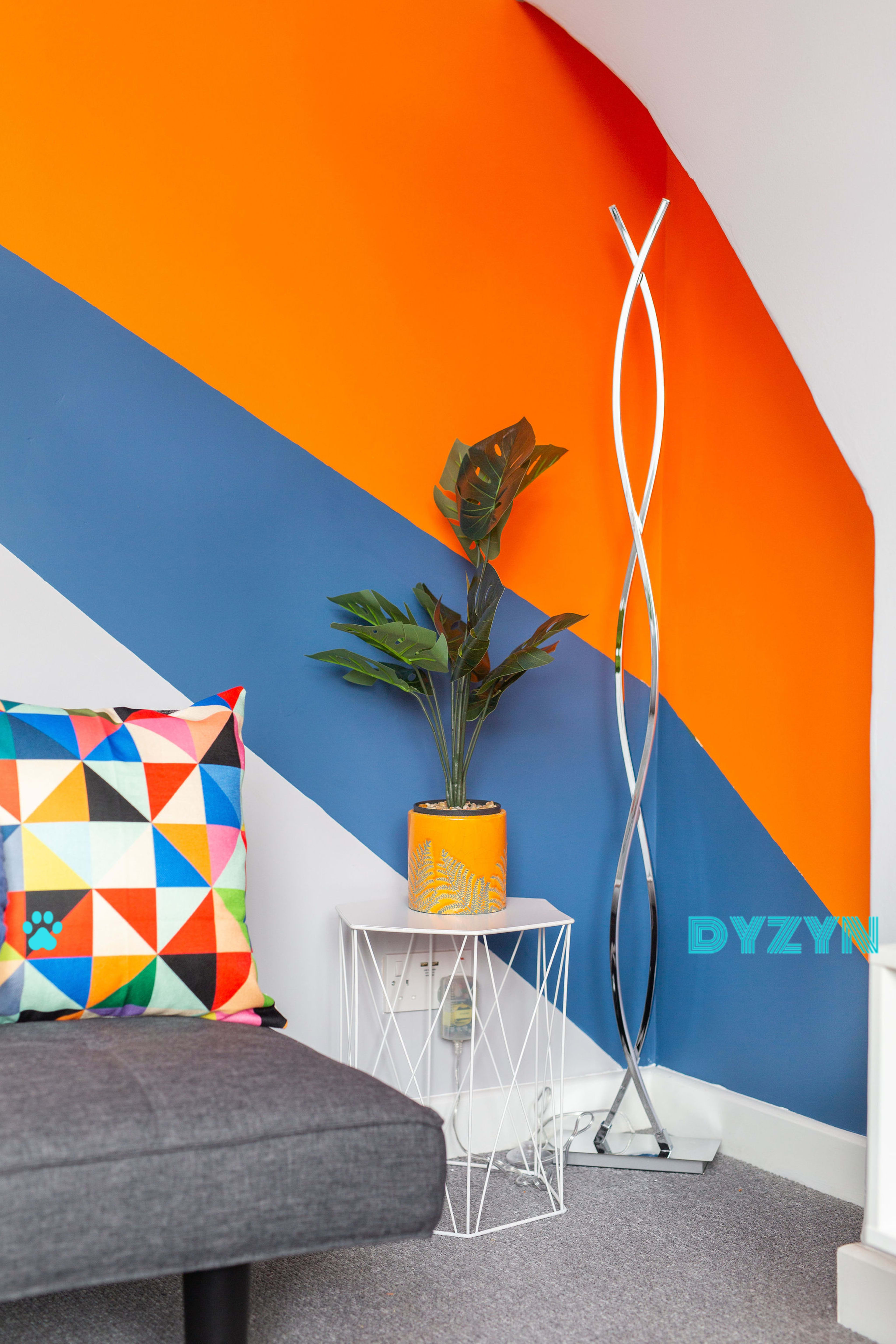 A corner of a room featuring an orange and blue accent wall, a geometric-patterned pillow on a gray sofa, a potted plant on a white side table, and a modern floor lamp.