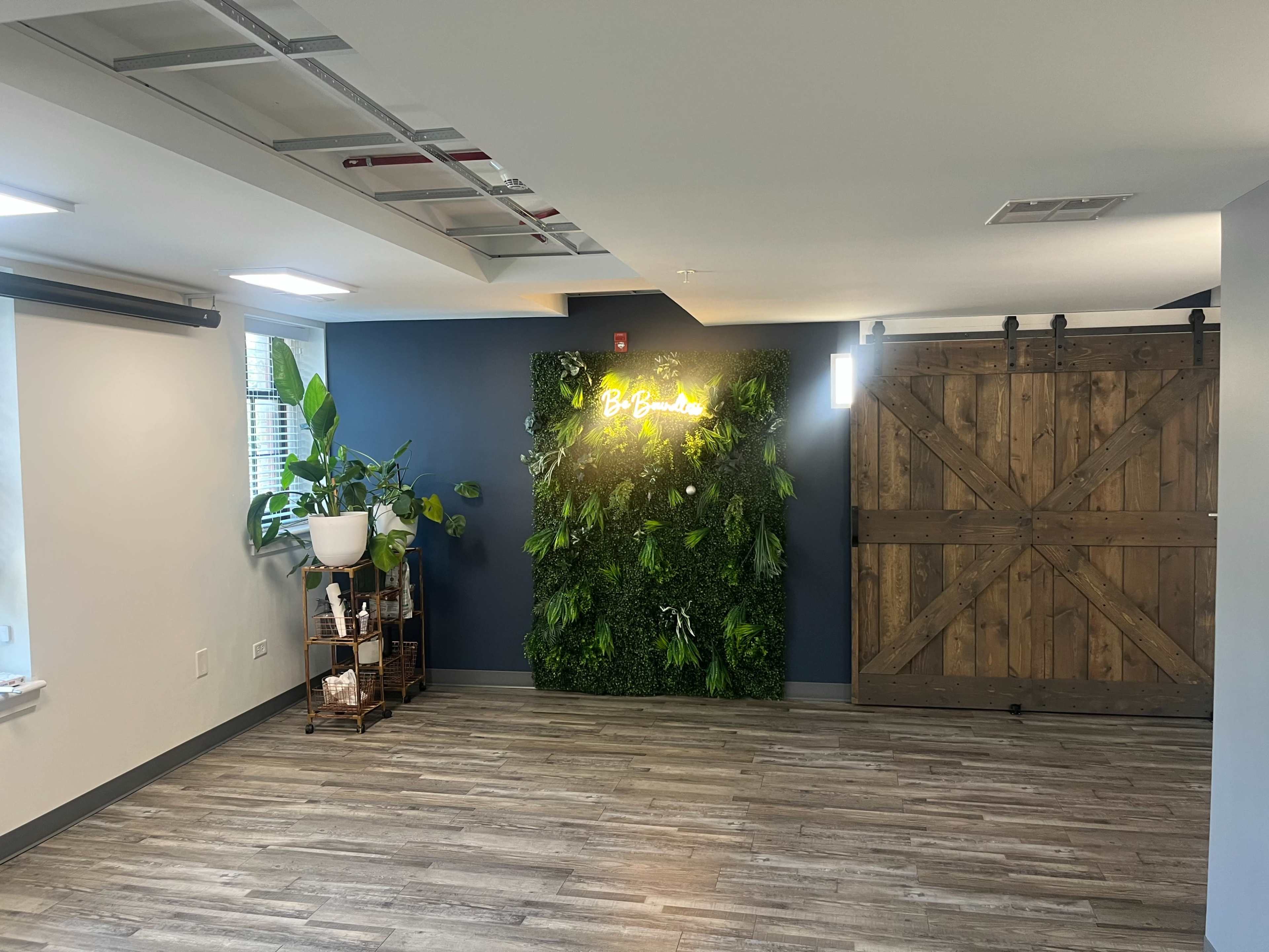 The room features a green wall with plants and a lighted sign, alongside a wooden sliding door and a small plant stand.