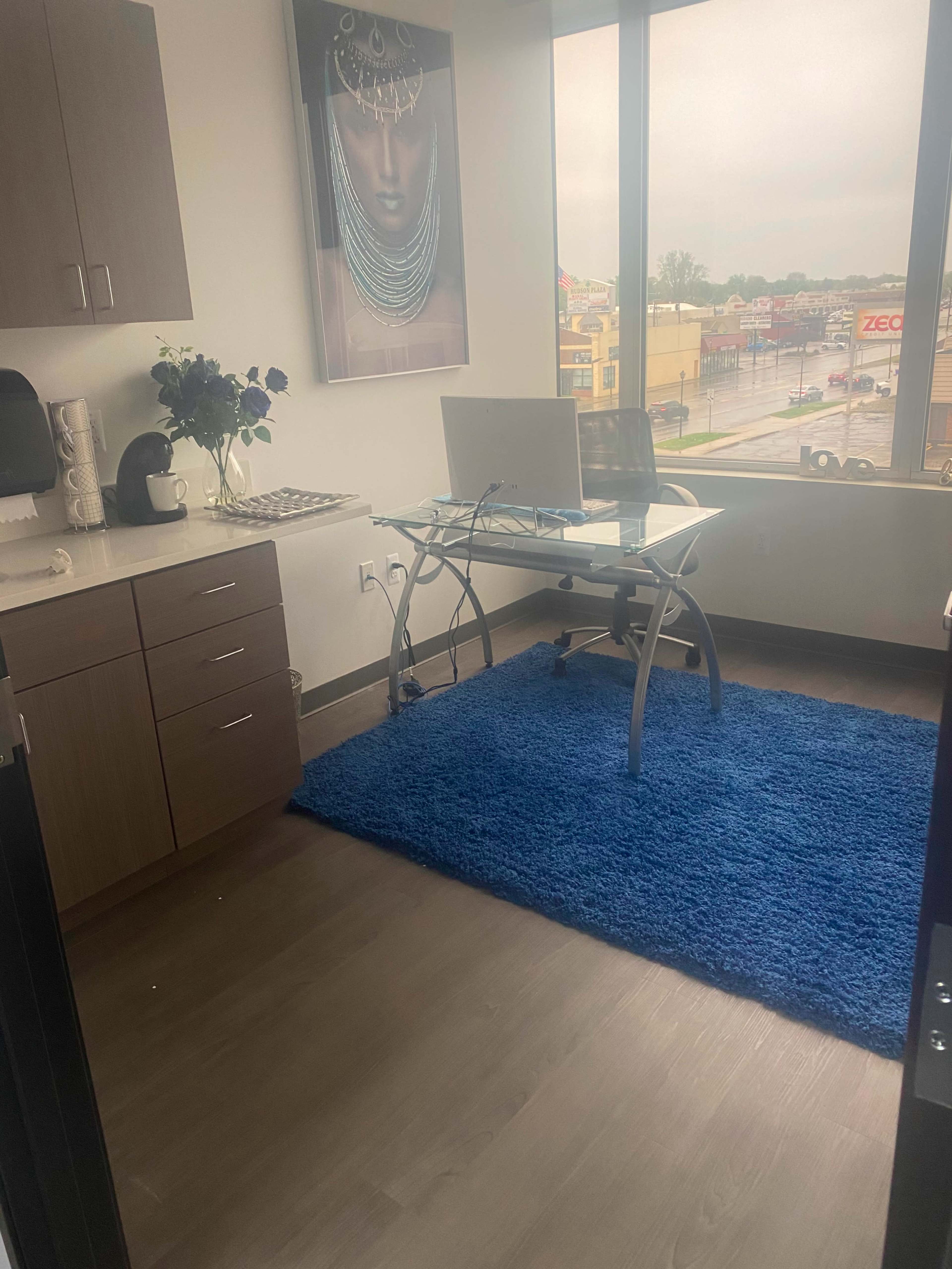 A modern office space features a glass desk with a computer, a blue rug, and large windows overlooking a street.