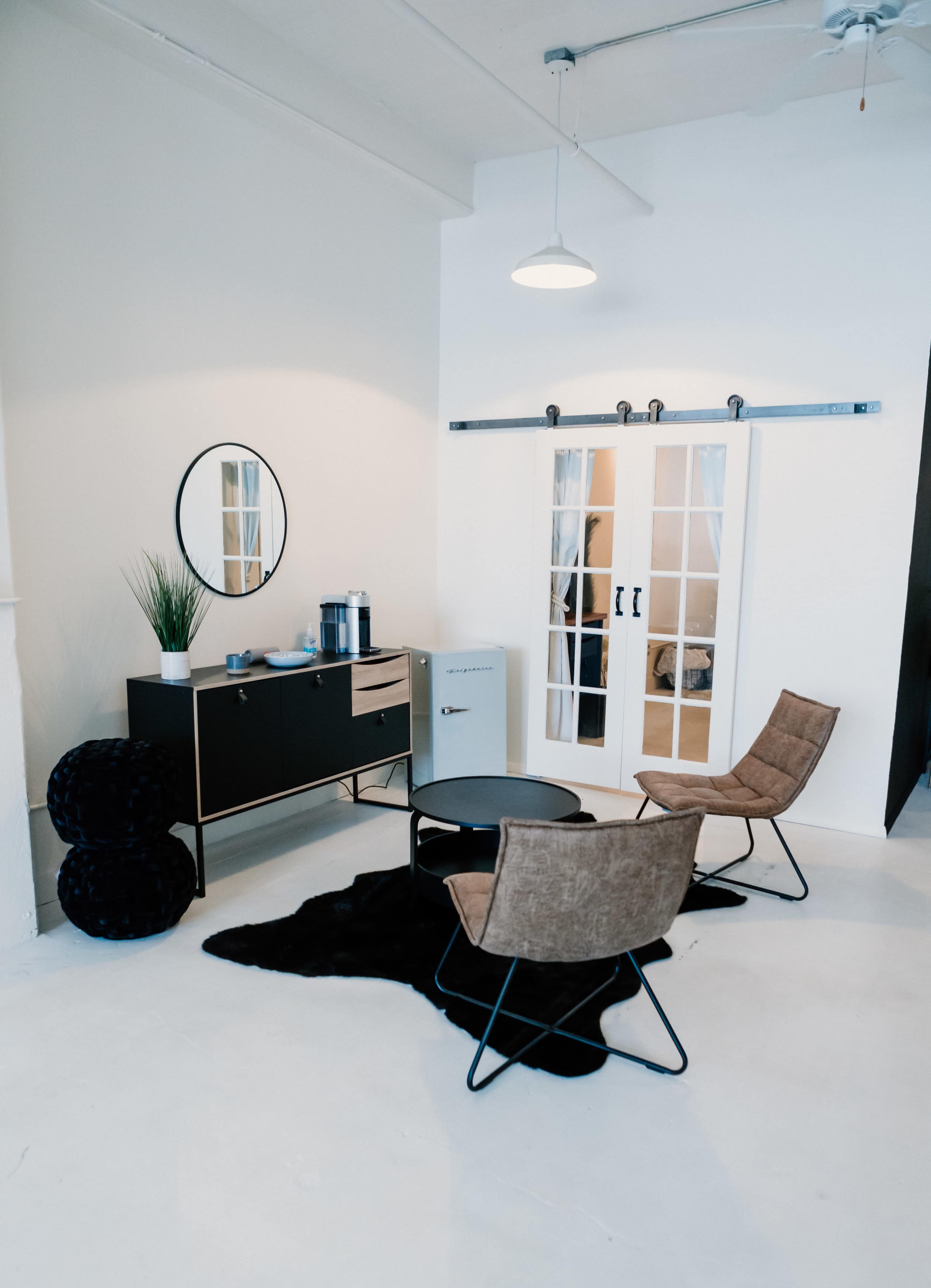 A modern minimalist living area featuring a coffee table, two upholstered chairs, a sideboard, a small plant, and a white refrigerator, with large sliding doors leading to another space.