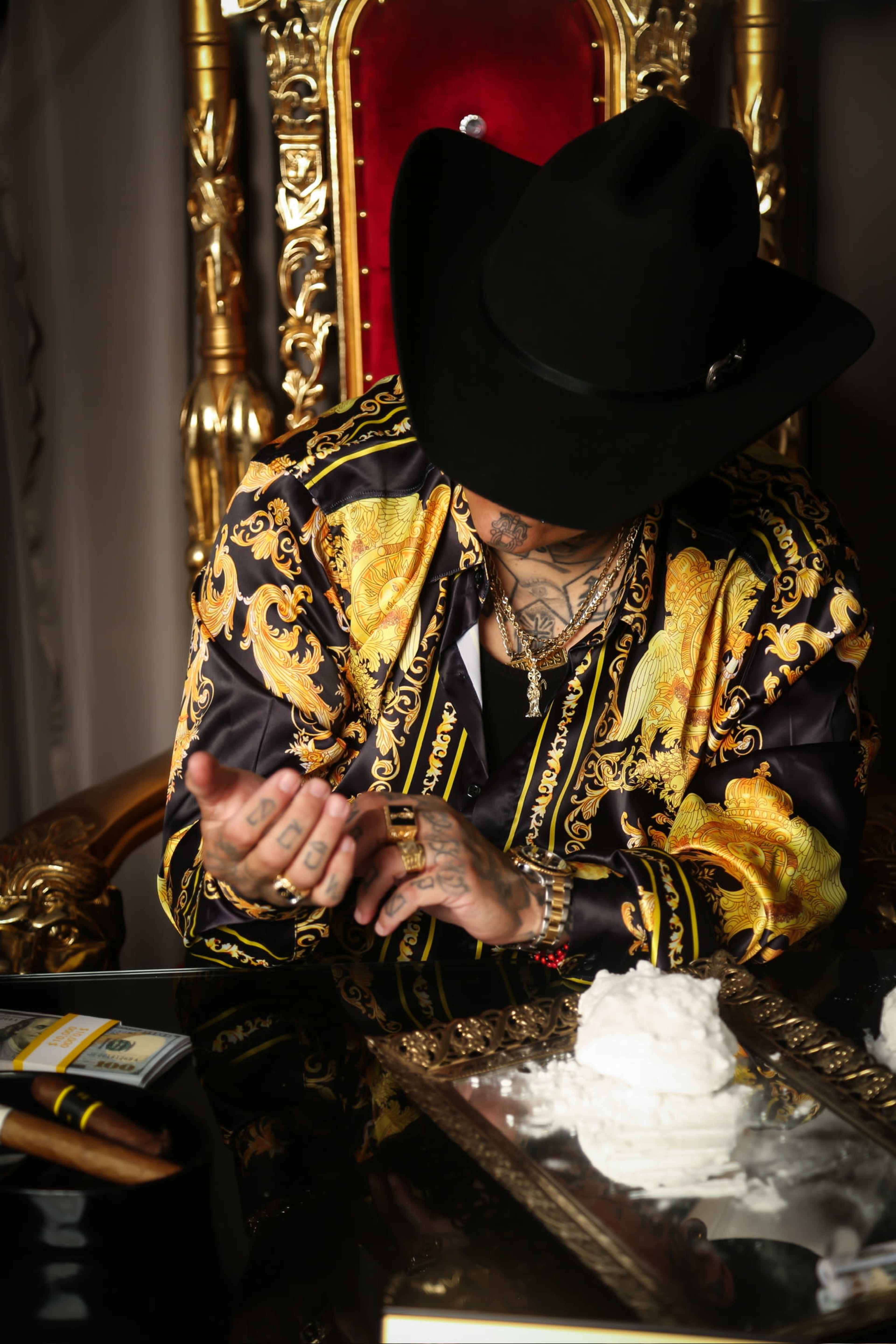 A person wearing a patterned shirt and a black hat sits on an ornate chair with a large mound of white powder and cash on a table in front of them.
