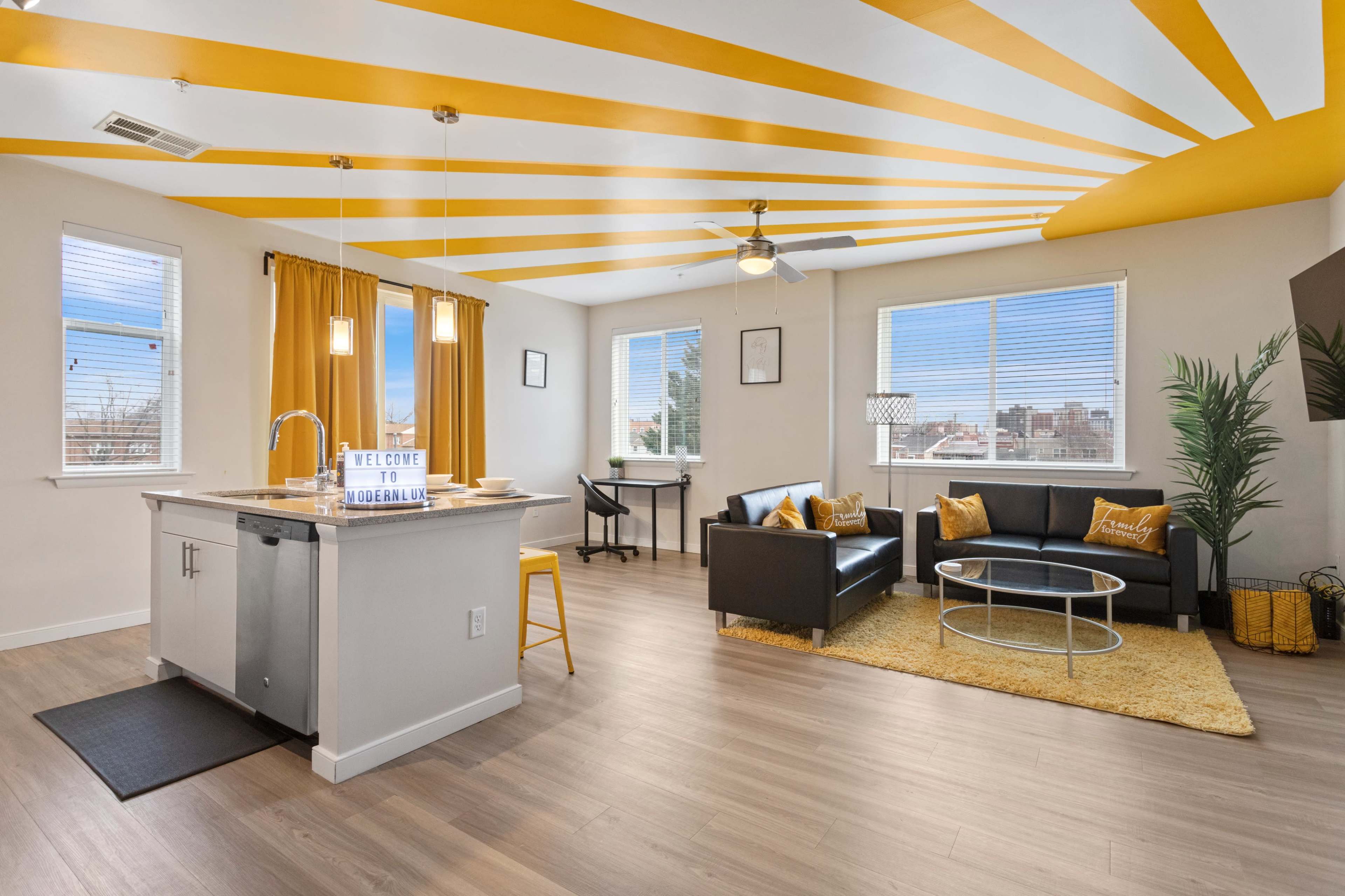 A bright living space features a kitchen area, a seating area with black furniture, and yellow accents, under a striped yellow and white ceiling.