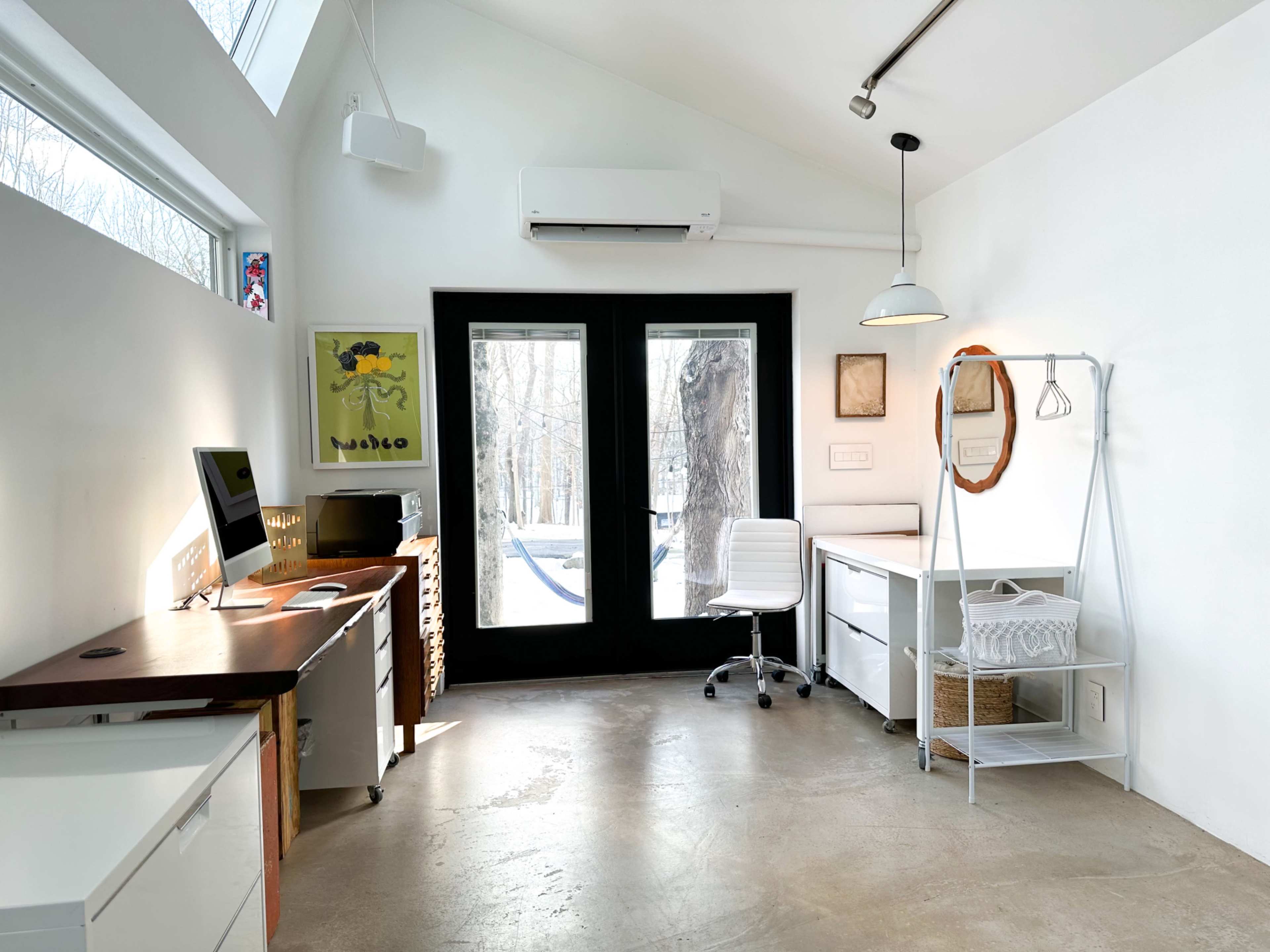 A bright, minimalist home office with large windows, a wooden desk, and a white chair, overlooking a wooded area.