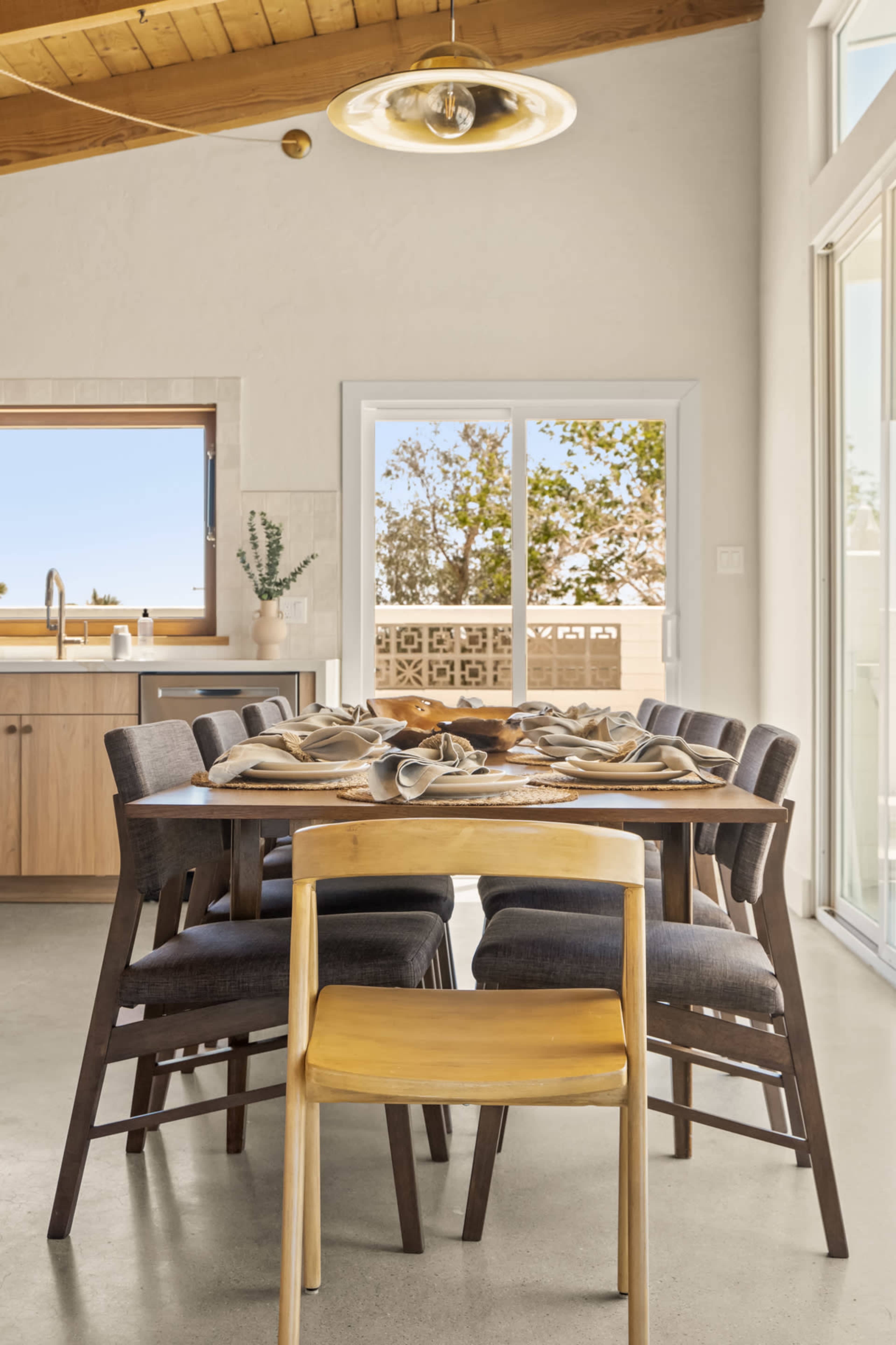 A dining table with eight chairs is set with place settings in a bright, modern kitchen space featuring large windows and wooden accents.