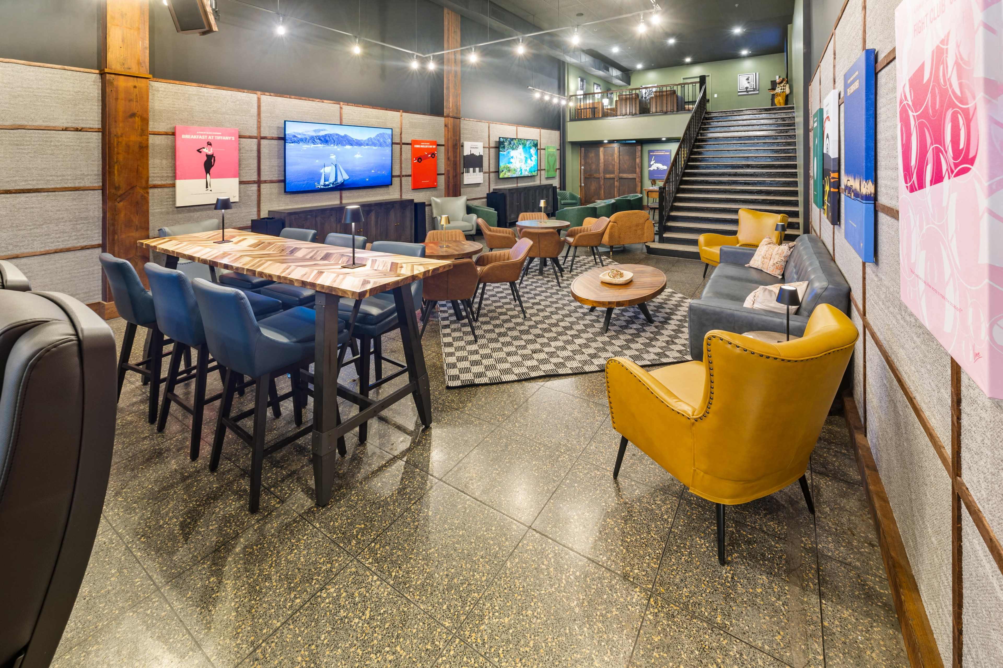The image shows a modern lounge area with a mix of table seating, armchairs, and a staircase, featuring art on the walls and large screens displaying various images.