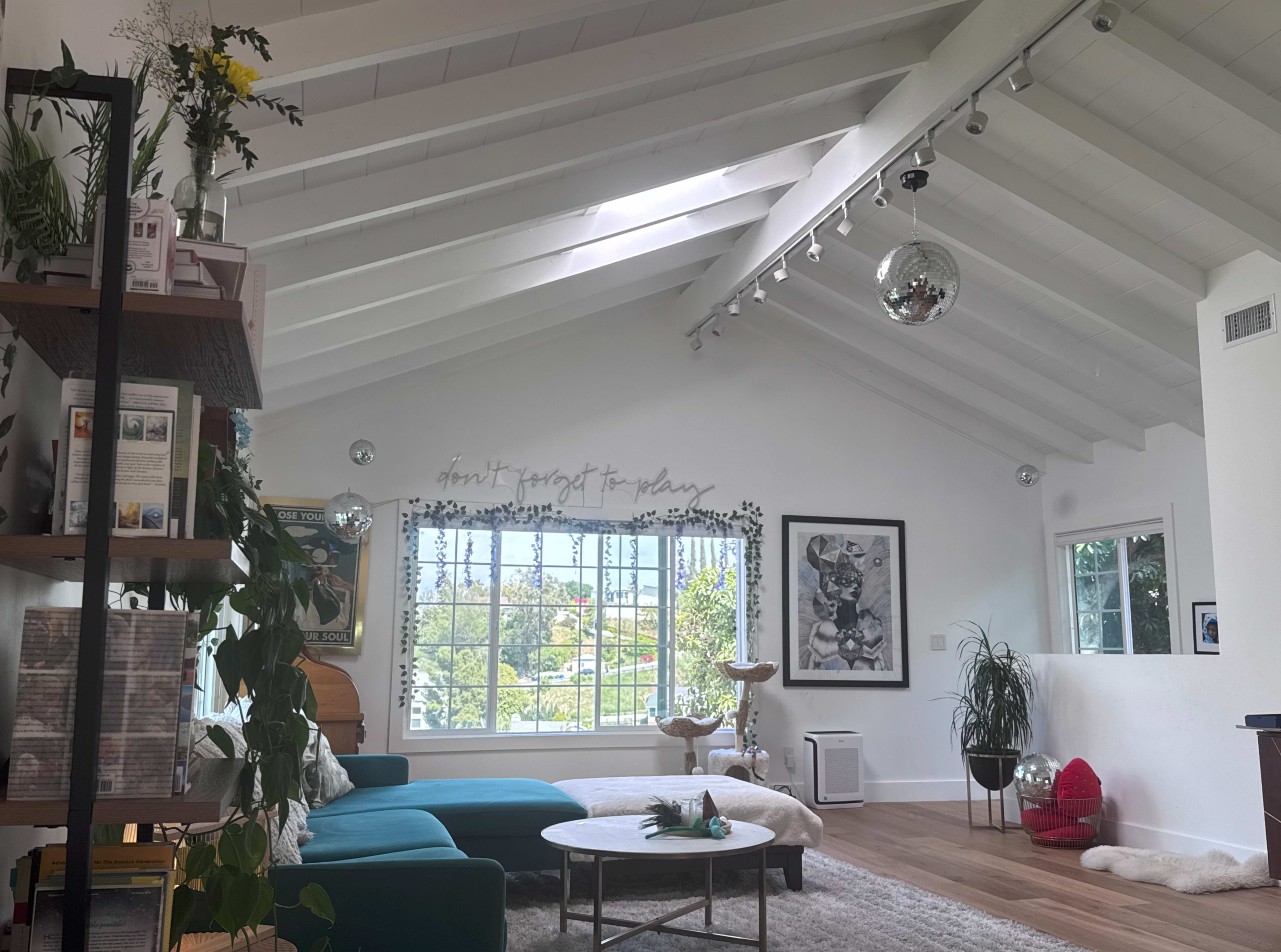 The image shows a bright living room with high, white-beamed ceilings, a large window, a teal couch, a round coffee table, and various plants.