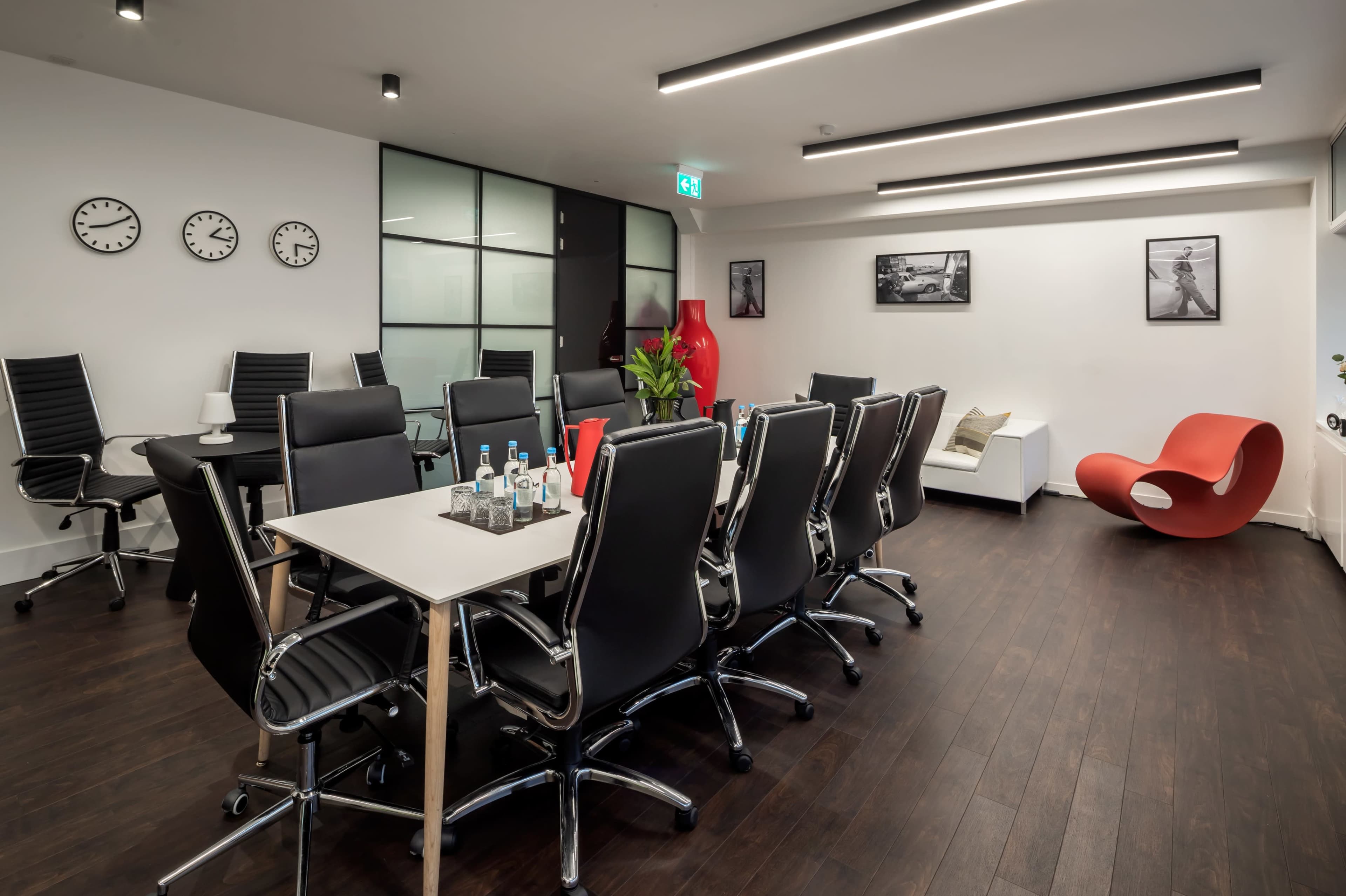 A modern conference room features a large table surrounded by black chairs, clocks on the wall, and a red sculpture in the corner.