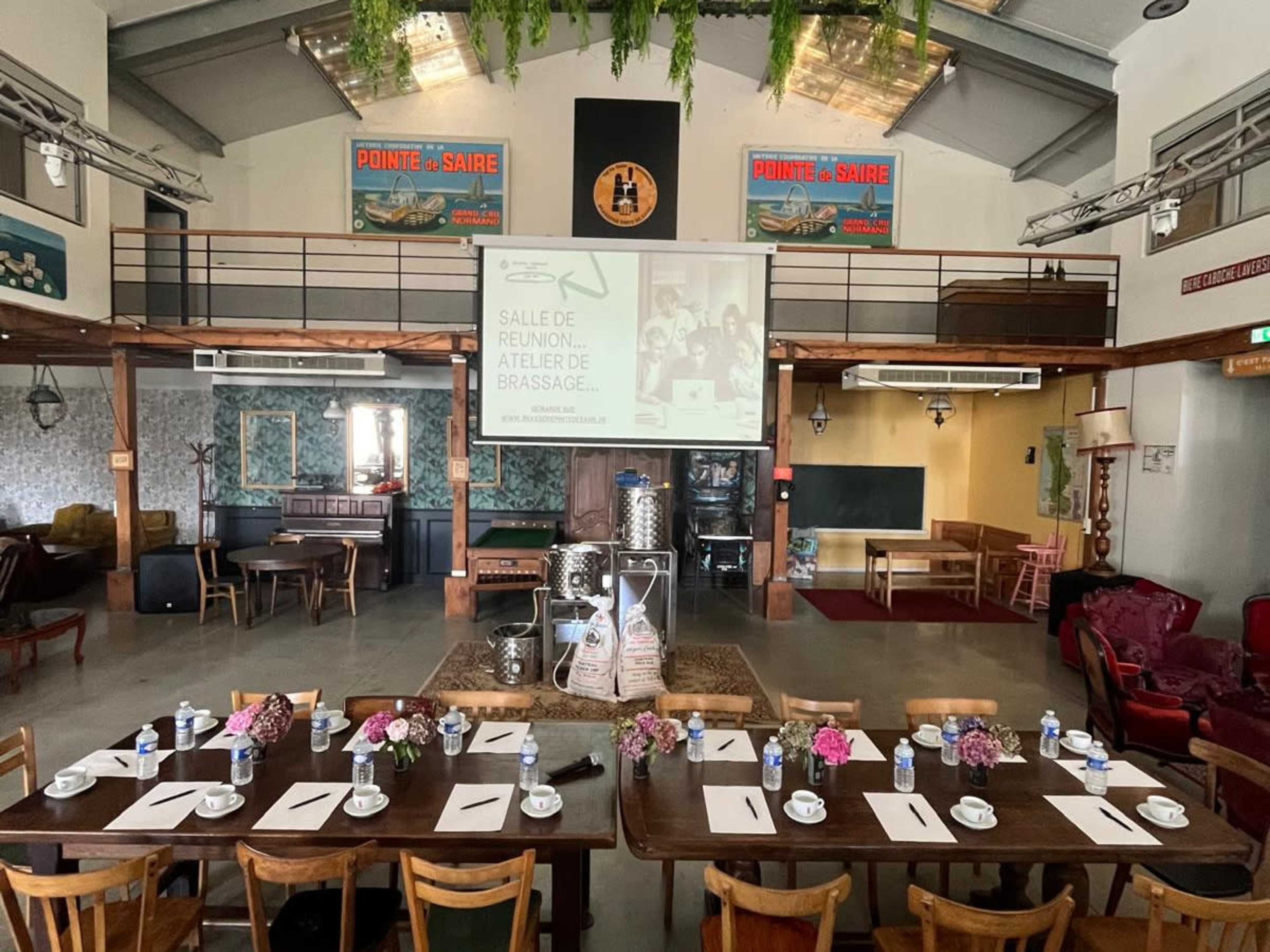 The image shows a spacious meeting room set up with a long table, floral centerpieces, and a projector screen at the front, surrounded by various seating areas and decorative elements.