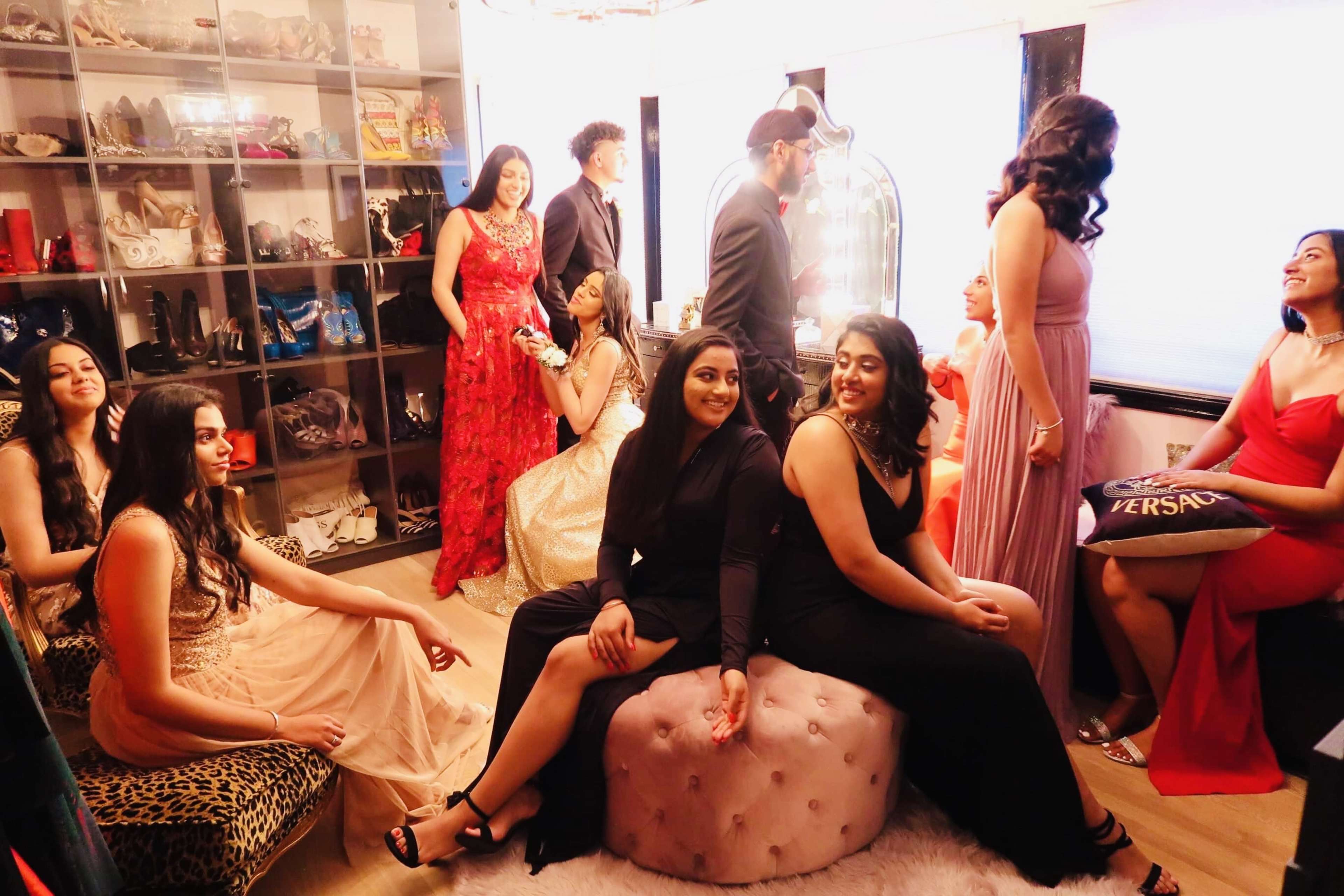 A group of young women dressed in evening gowns and formal attire socialize in a stylish room filled with shoes and accessories.