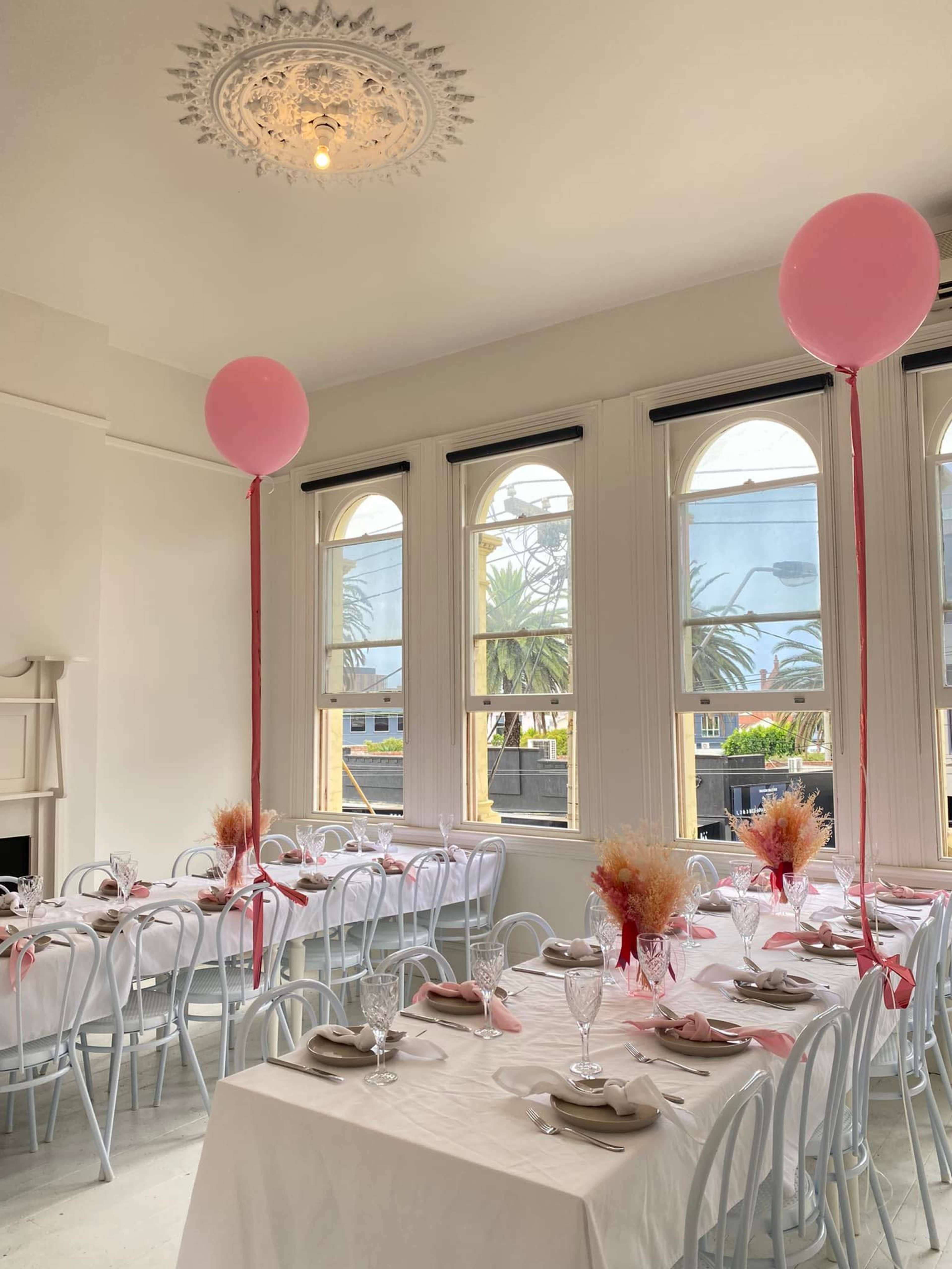 A dining area features white tables set with beige napkins and decorated with pink balloons and floral centerpieces under large arched windows.