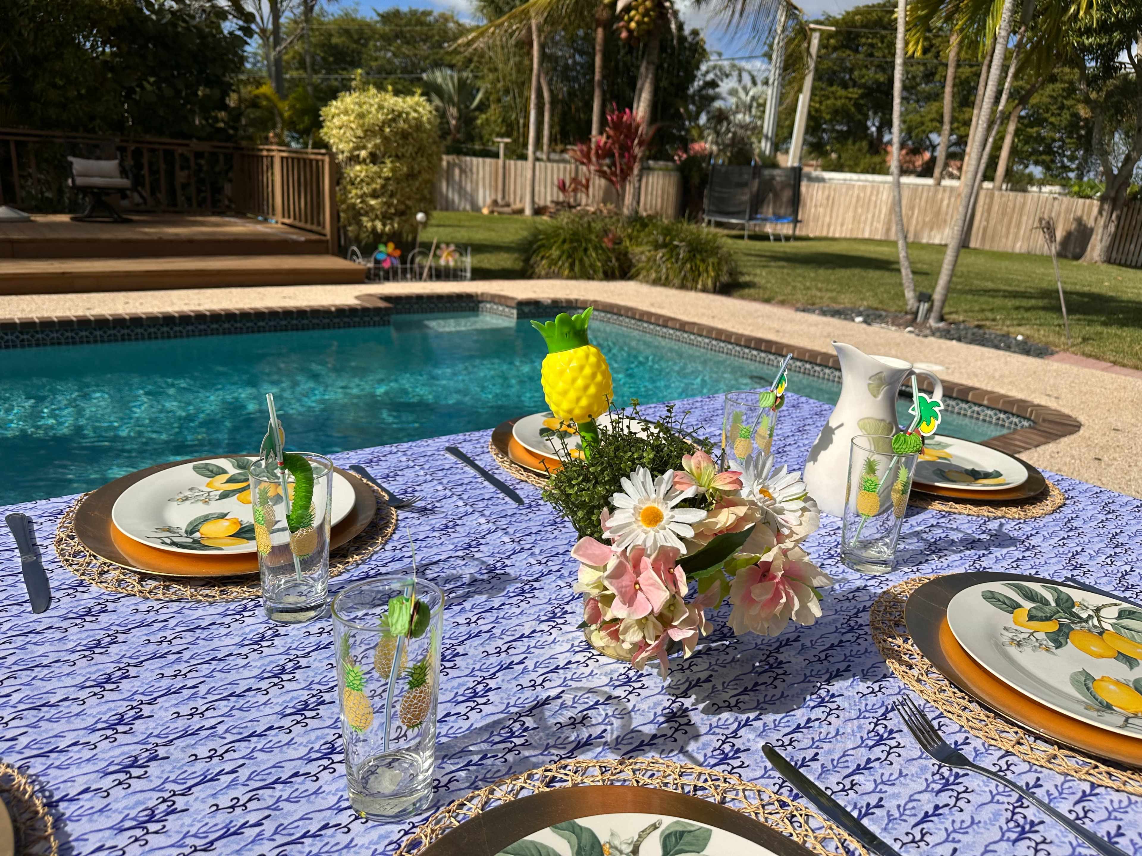 A dining table is set with decorative plates and glasses beside a swimming pool surrounded by tropical plants.