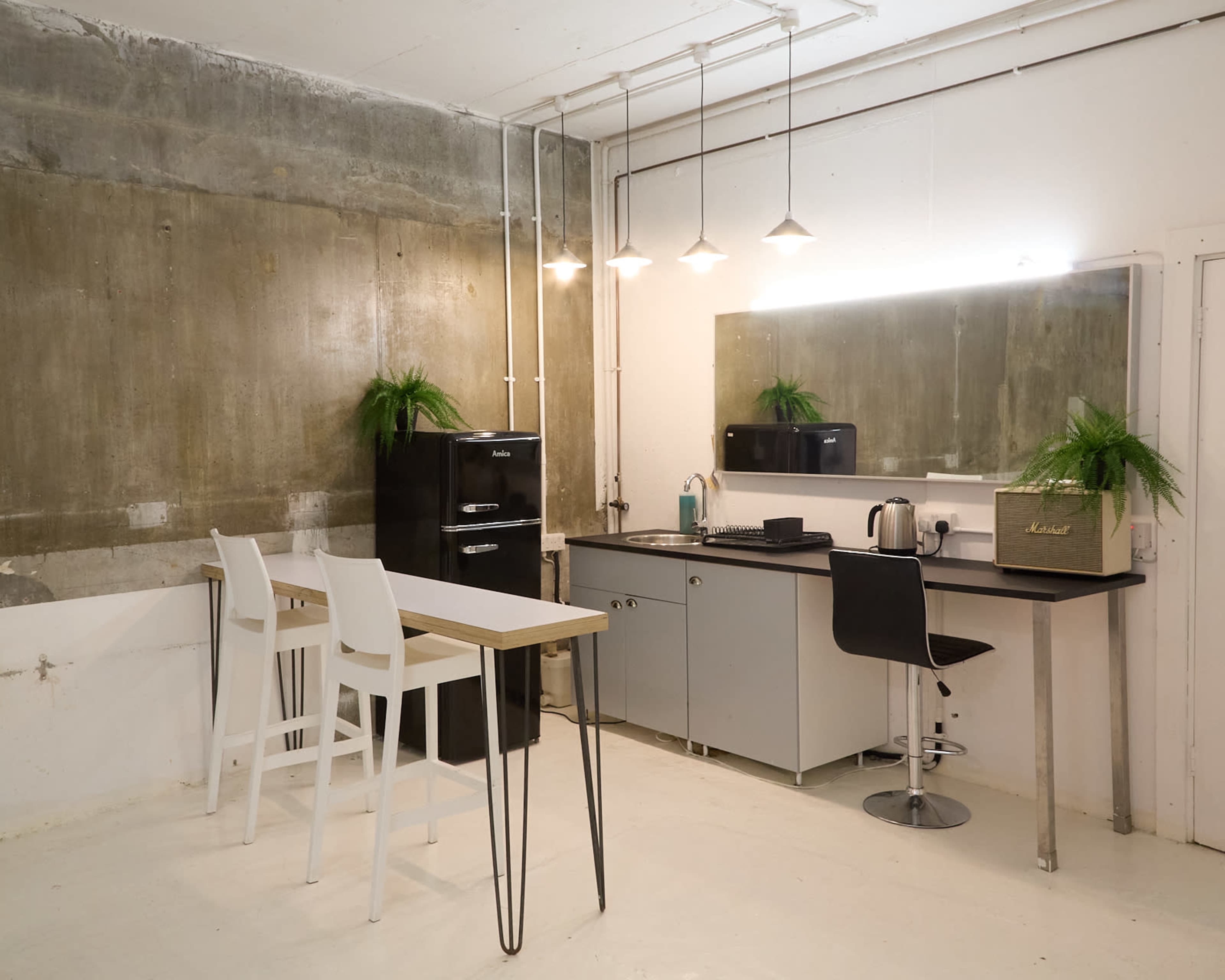 The image shows a modern kitchenette with a black refrigerator, a sink, a kettle, and two seating areas, set against a partially exposed concrete wall.
