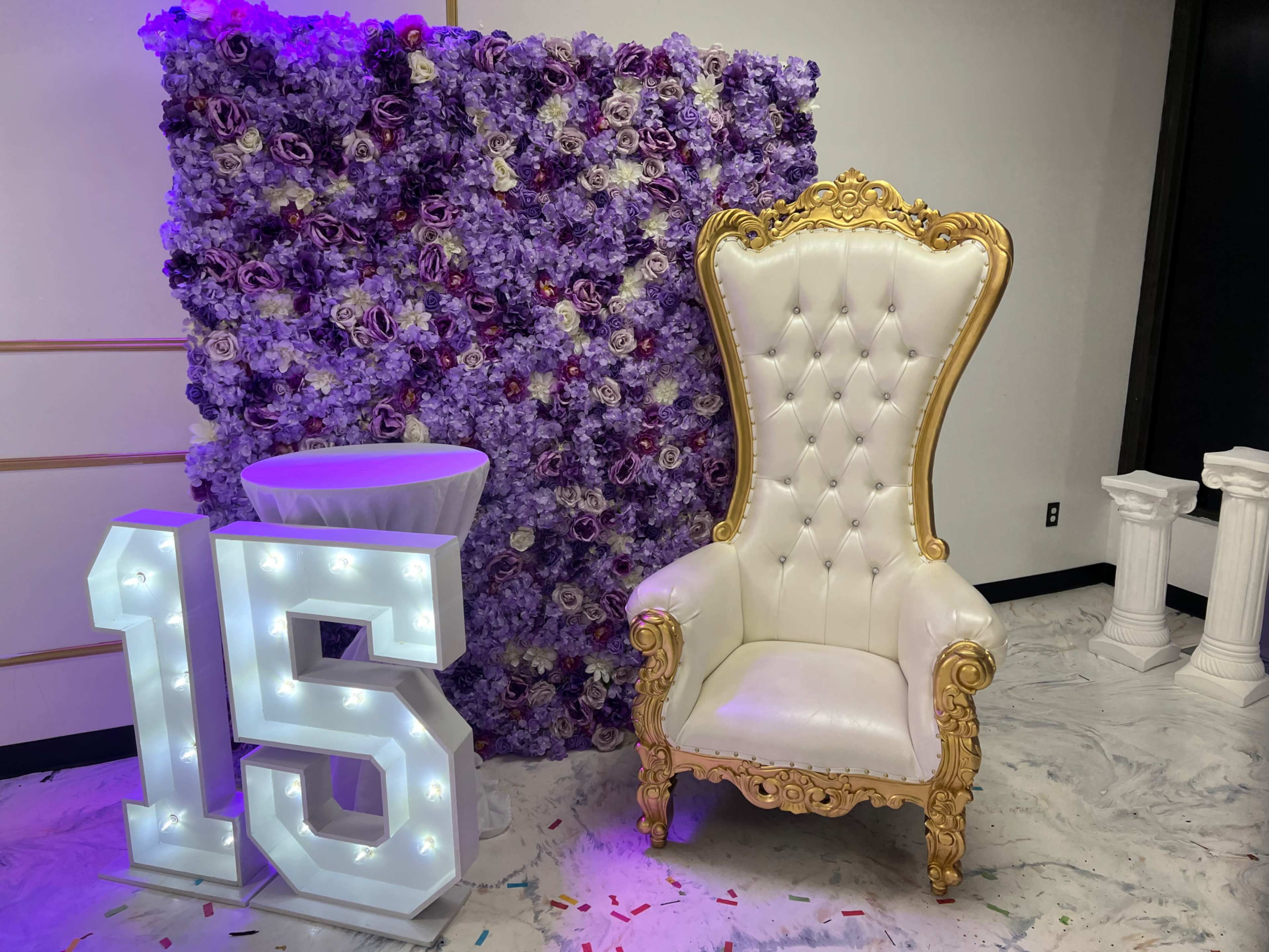 The image shows a decorative setup for a celebration, featuring a large white and gold throne chair, a purple floral backdrop, a white table, and illuminated numbers "15."