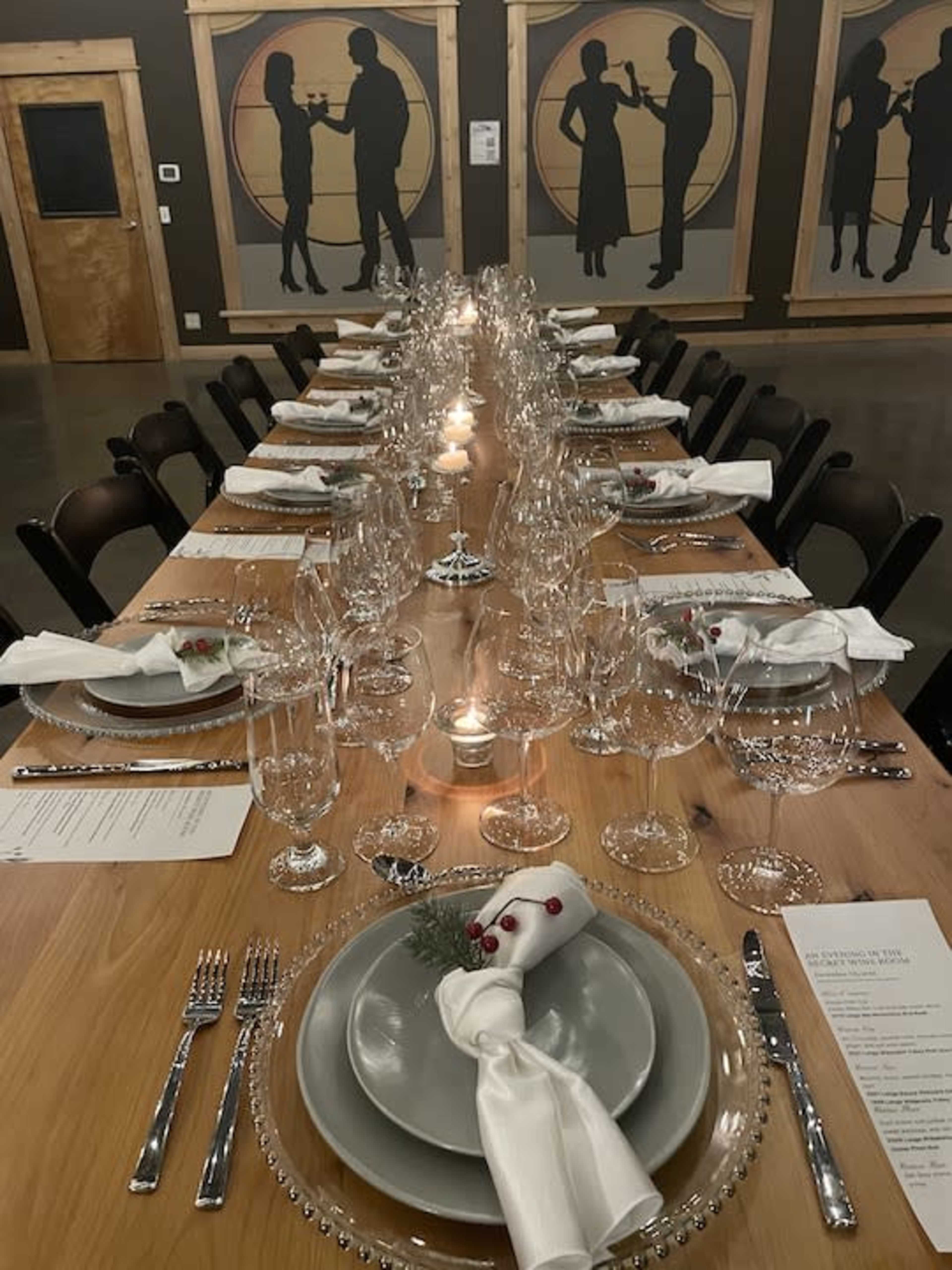 A long wooden dining table is elegantly set with fine china, glassware, and white napkins, surrounded by chairs in a room featuring silhouette artwork on the walls.