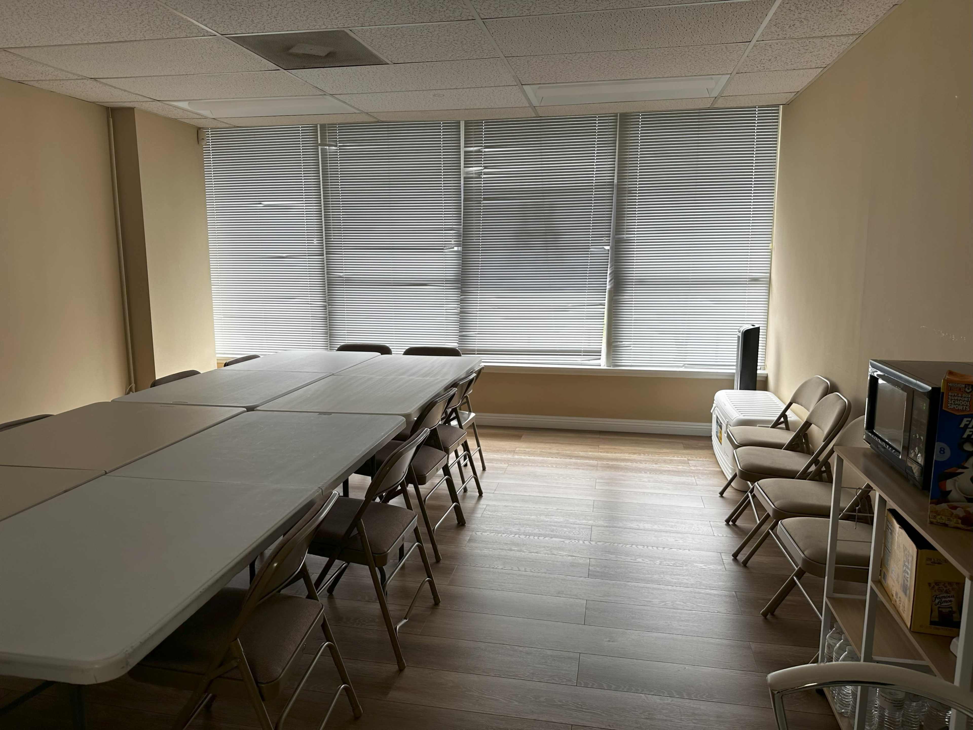 A meeting room features a long table surrounded by folding chairs and large windows with blinds.