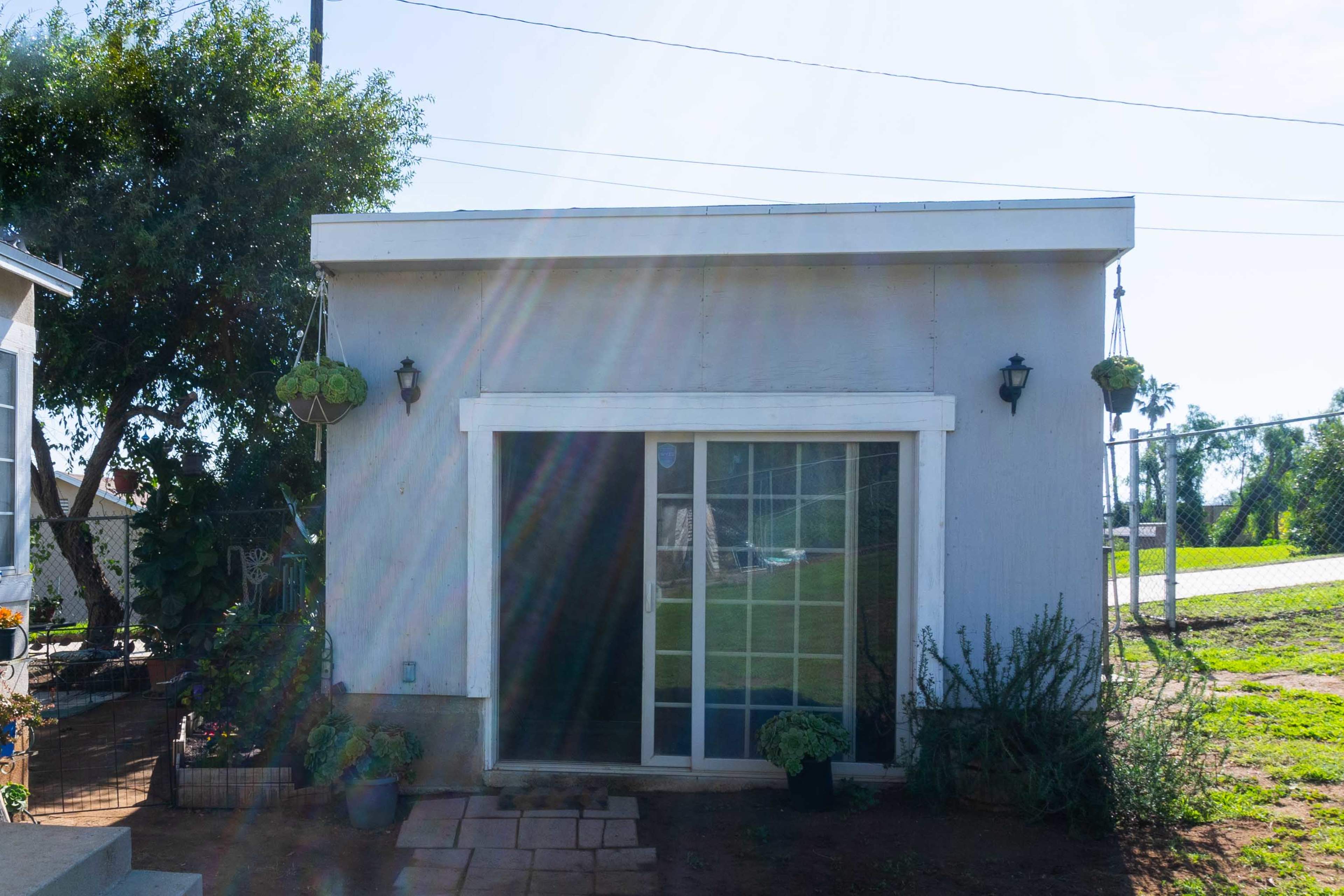 A small gray building with a sliding glass door is surrounded by plants and has two wall-mounted lights on its exterior.
