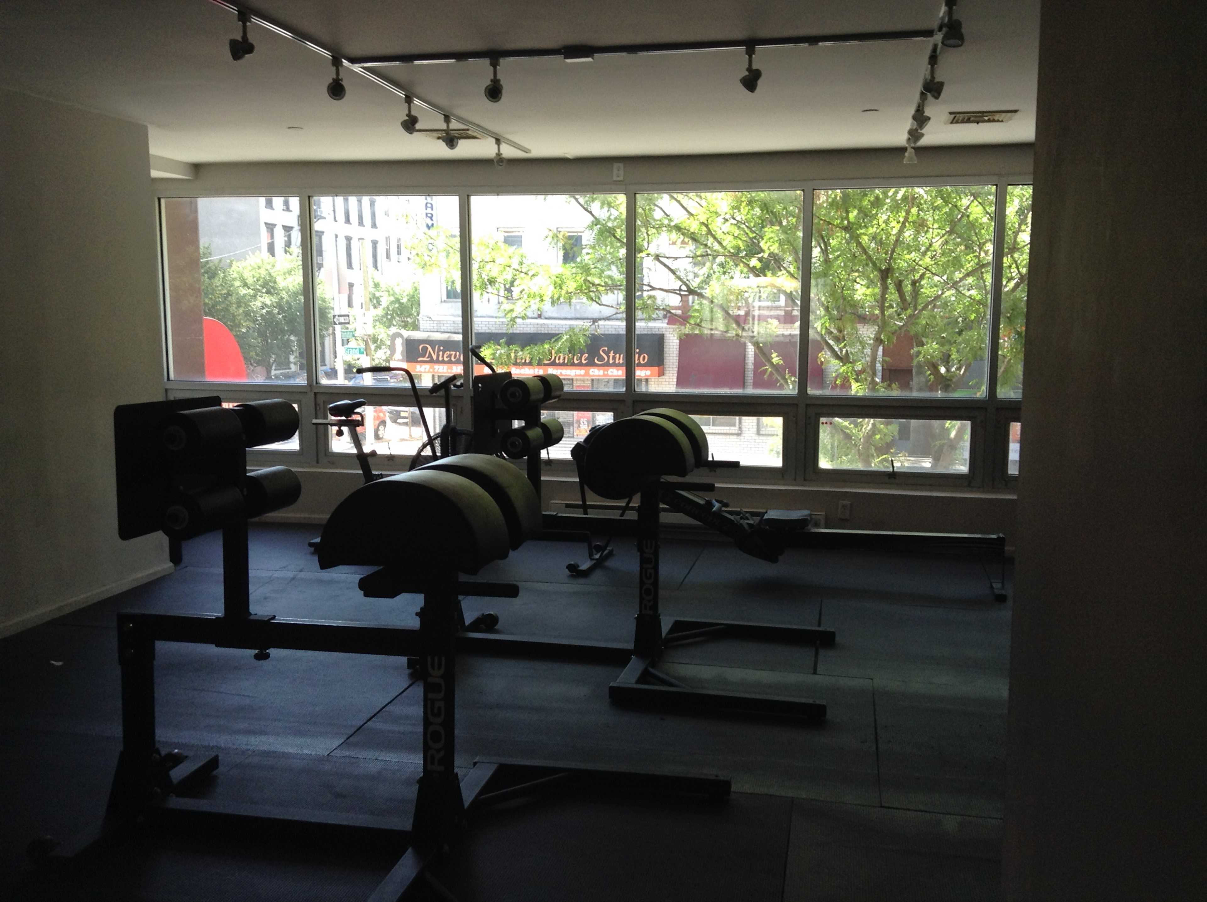 A gym interior with fitness equipment, including weight benches and machines, under natural light from large windows.