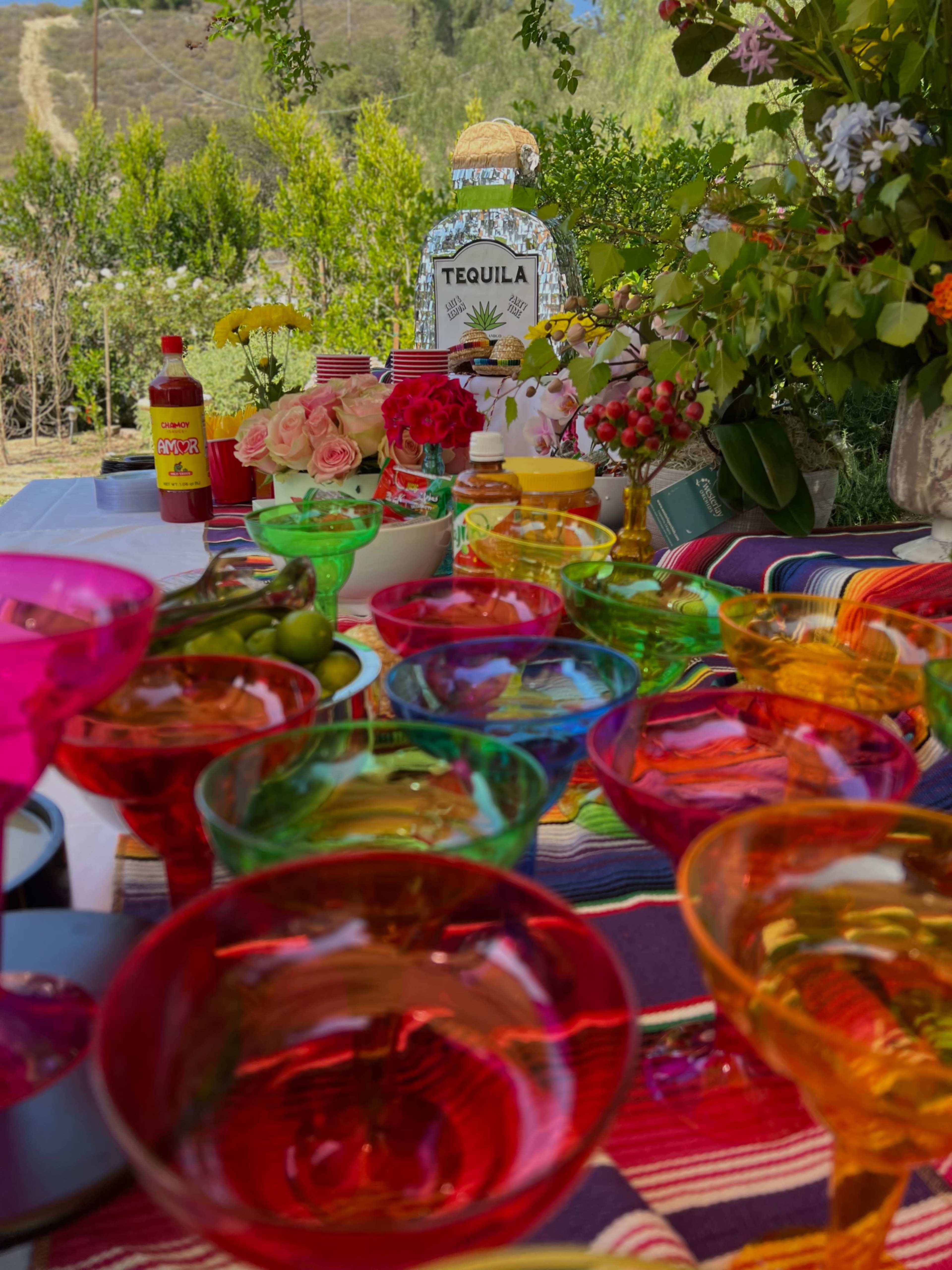 A vibrant table is set with colorful cocktail glasses, a bottle of tequila, and decorative flowers against a backdrop of greenery.