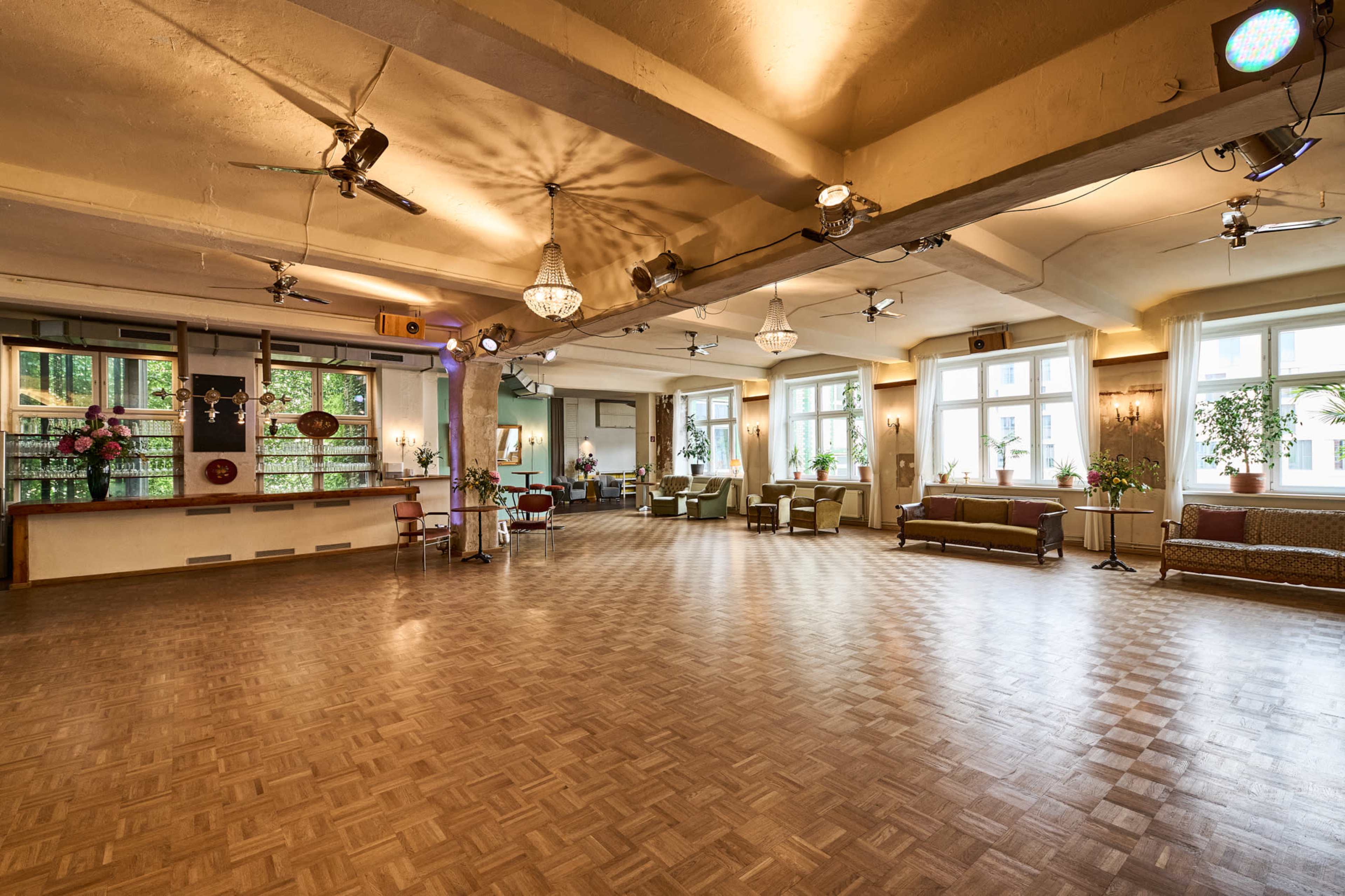 An empty event space with wooden flooring, large windows, and scattered seating arrangements.