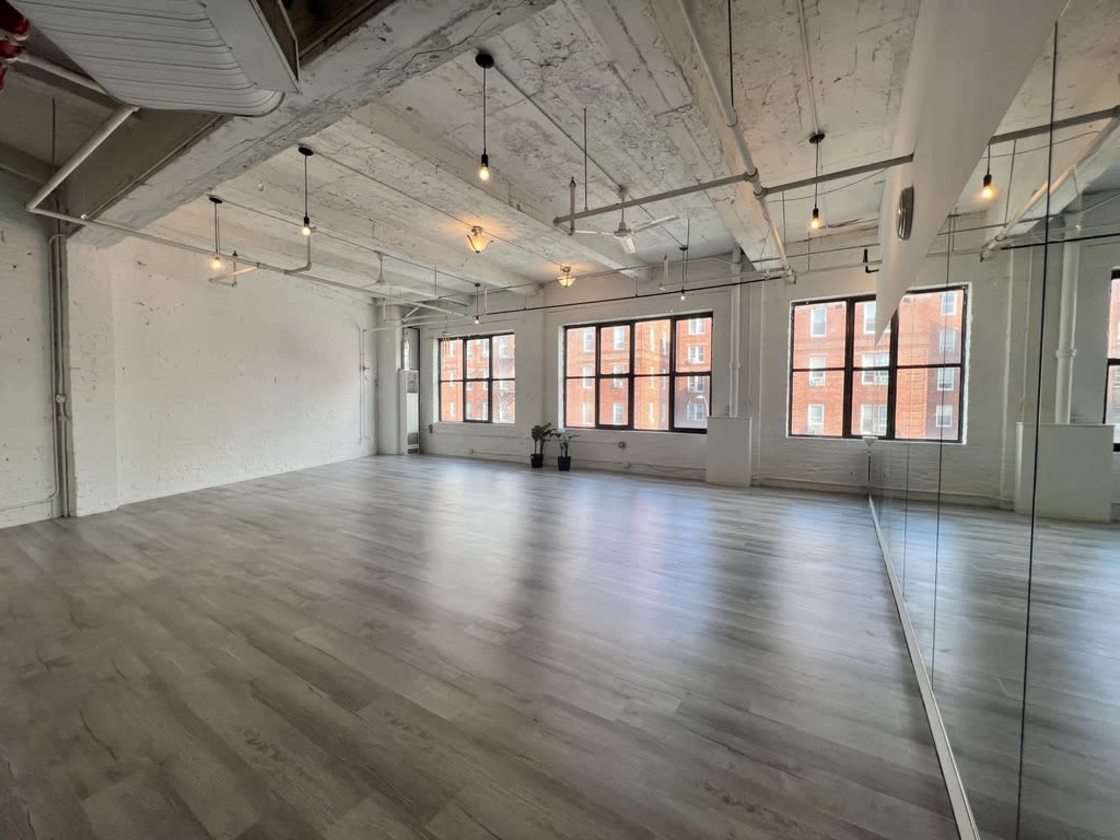 The image shows a spacious, minimalist room with large windows, wooden floors, and exposed ceilings, featuring a mirrored wall.