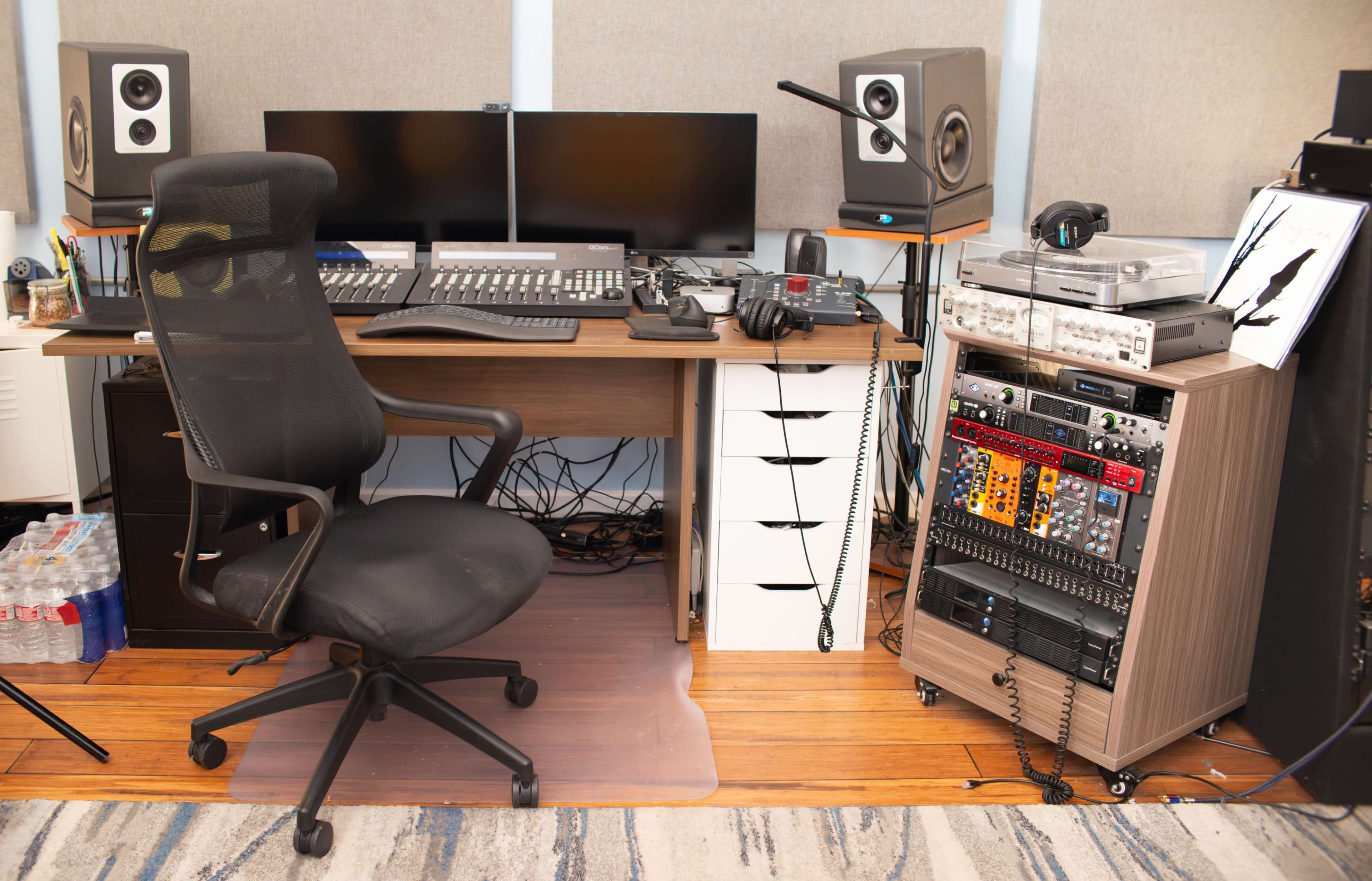 The image shows a music production studio featuring a computer desk with dual monitors, audio equipment, and various musical instruments, alongside a recording rack and a black office chair.