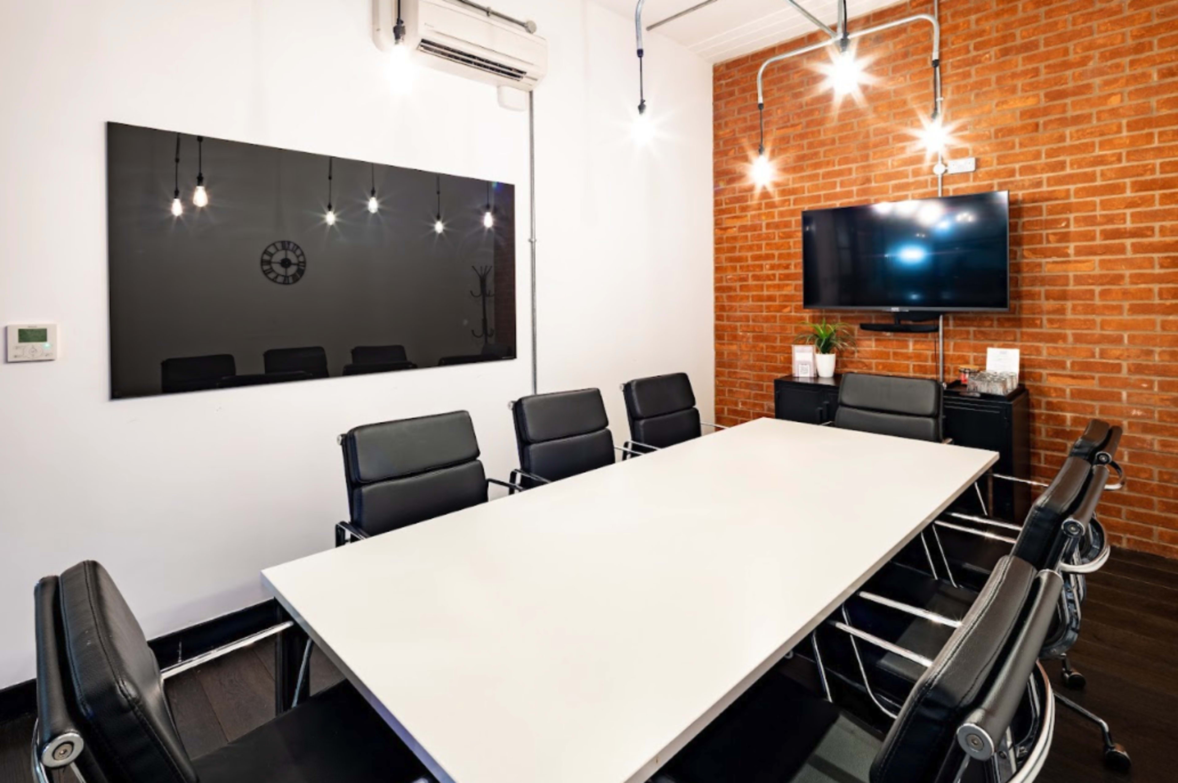 A modern conference room features a long white table surrounded by black chairs, with a brick accent wall and two screens mounted on opposite walls.