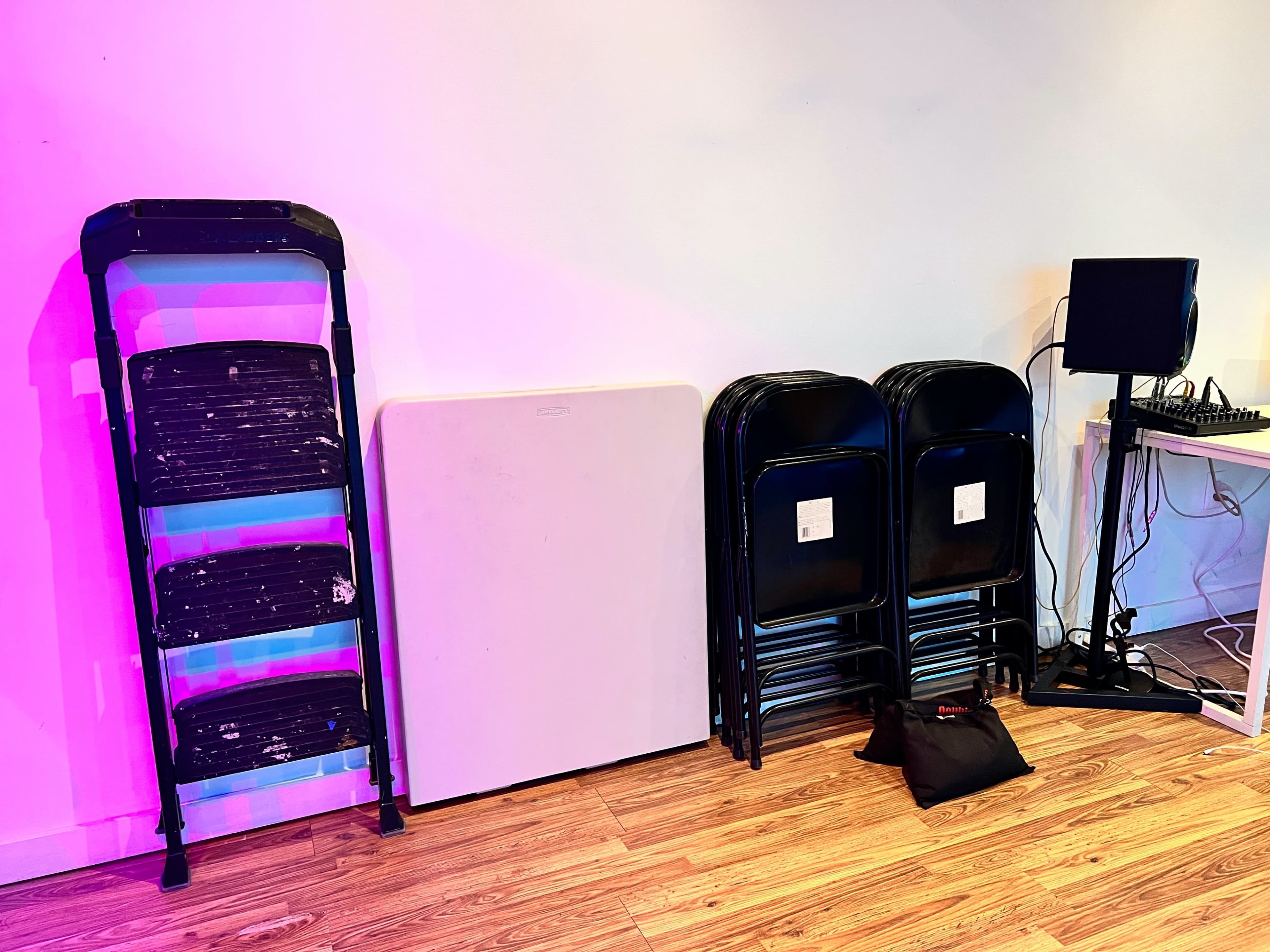 The image shows a stack of black folding chairs, a white folding table, and a speaker next to a sound mixing board against a wall with purple lighting.