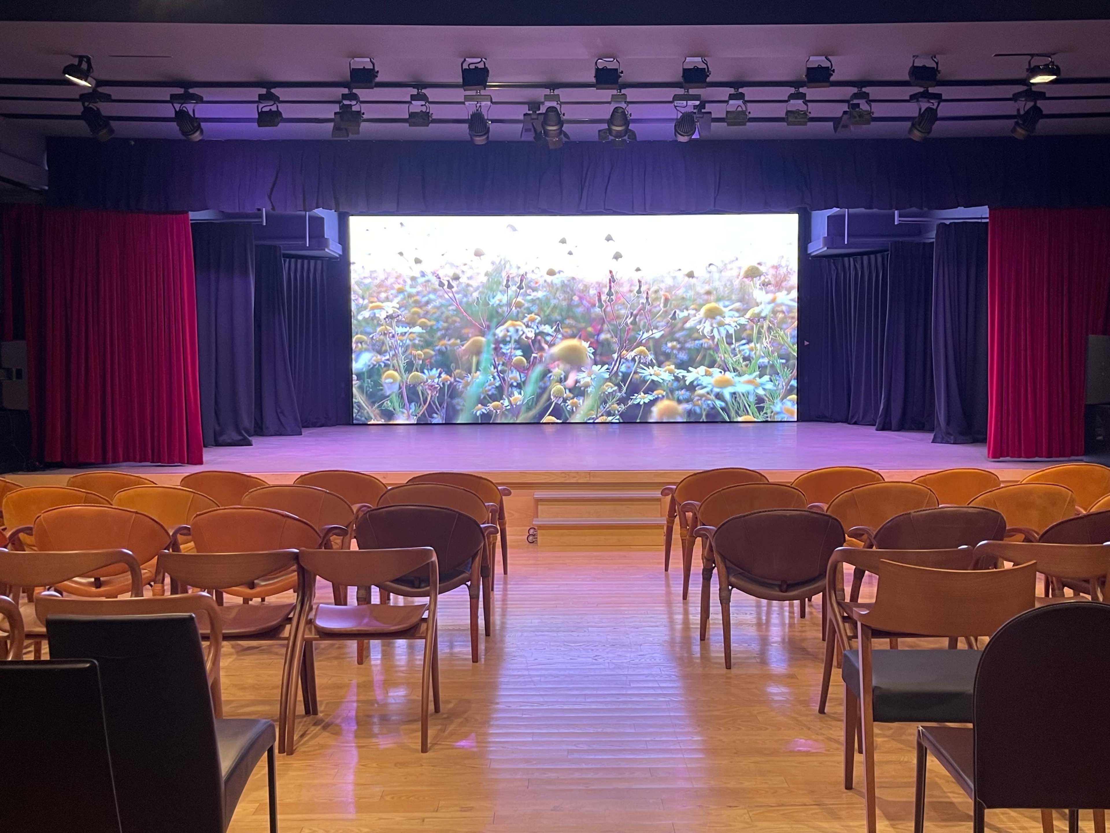 The image shows a stage with a large screen displaying a floral scene, set up in front of a row of wooden chairs in an auditorium.