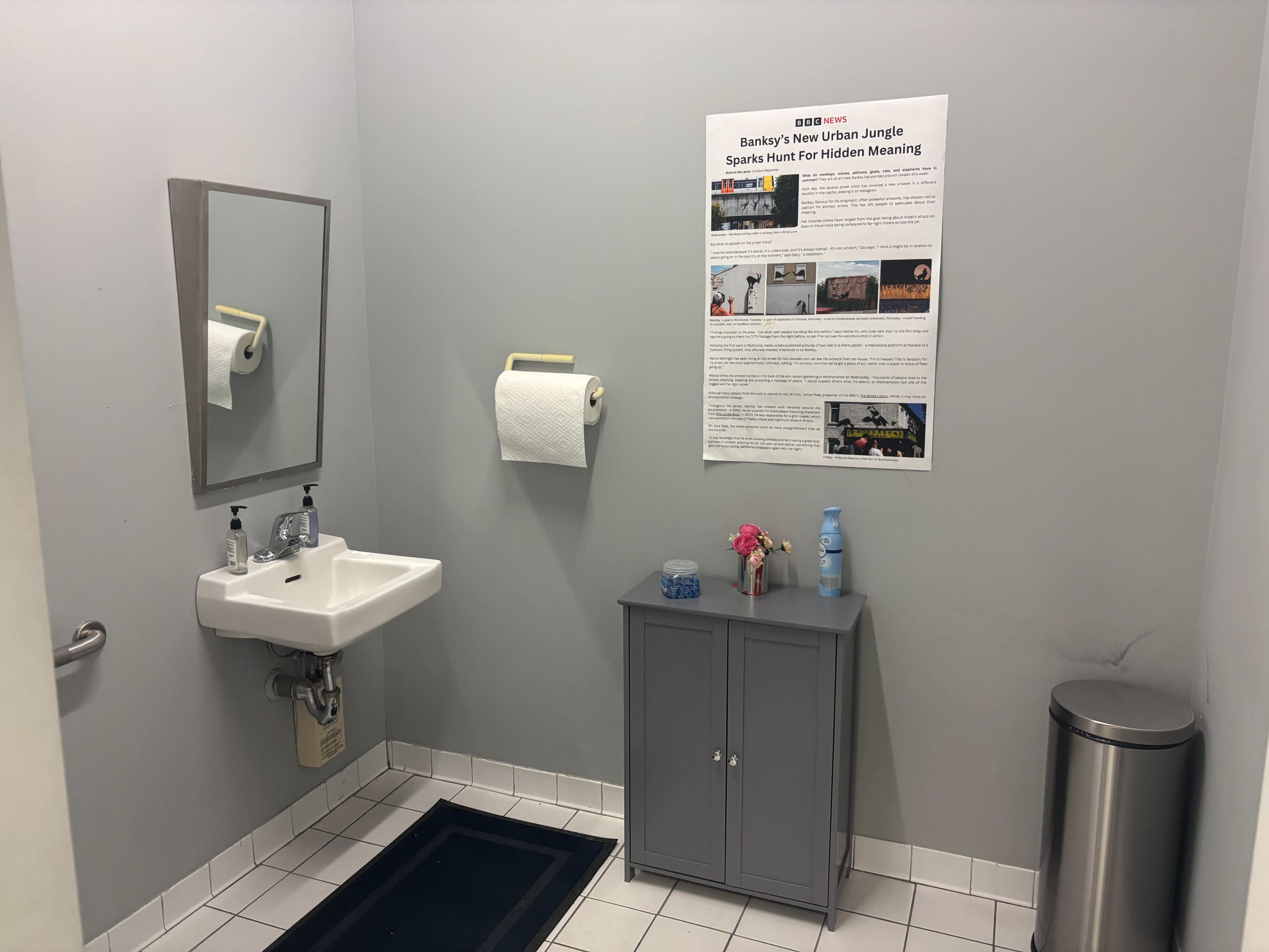 The image shows a small bathroom with a sink, a mirror, a paper towel holder, a poster on the wall, a cabinet under the sink, a trash can, and a dark mat on the floor.