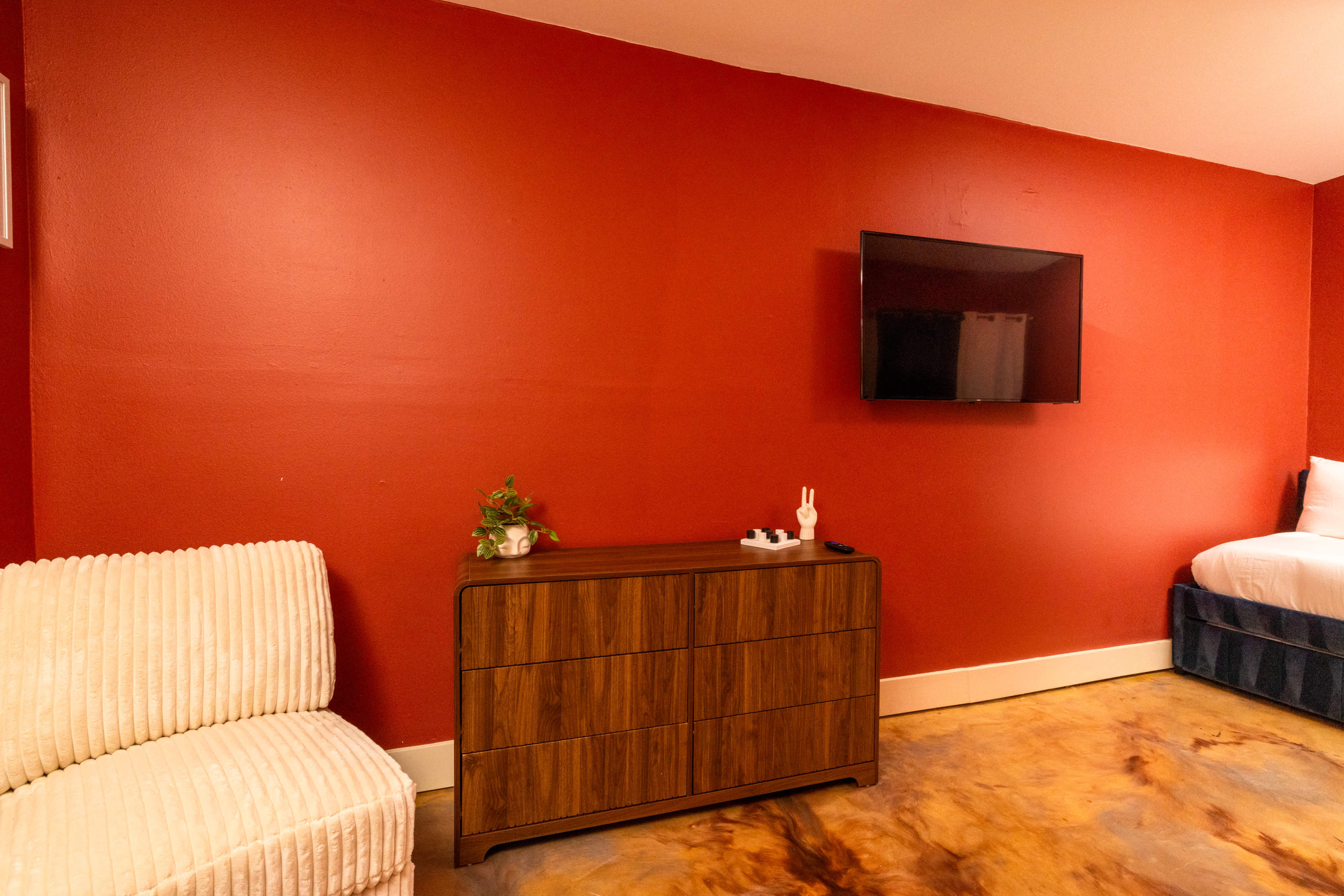 The image shows a room with a red wall, a television mounted above a wooden dresser, a white plush chair, and a bed partially visible in the background.
