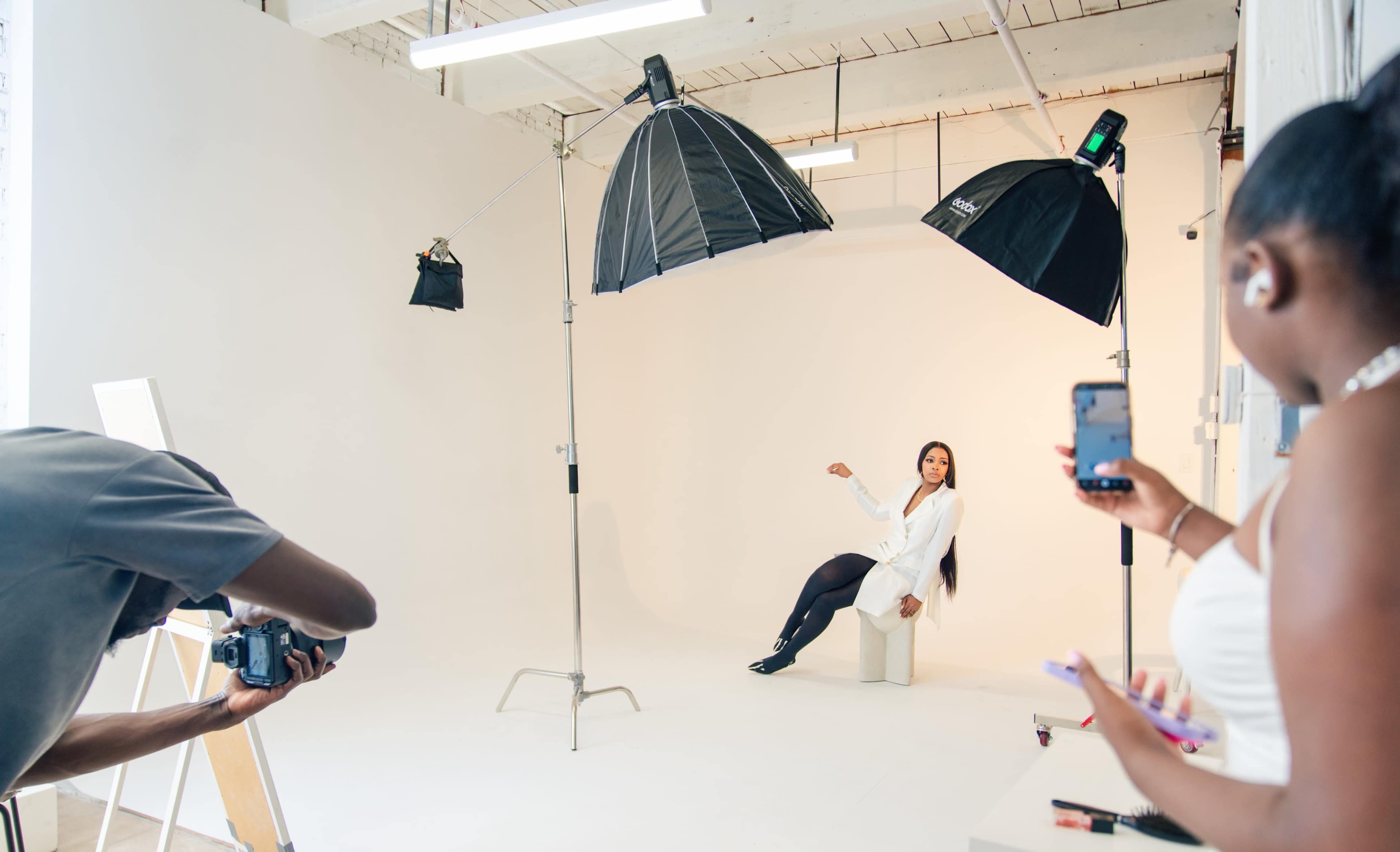 A model poses on a stool in a well-lit photography studio while a photographer takes pictures and another person uses a smartphone.