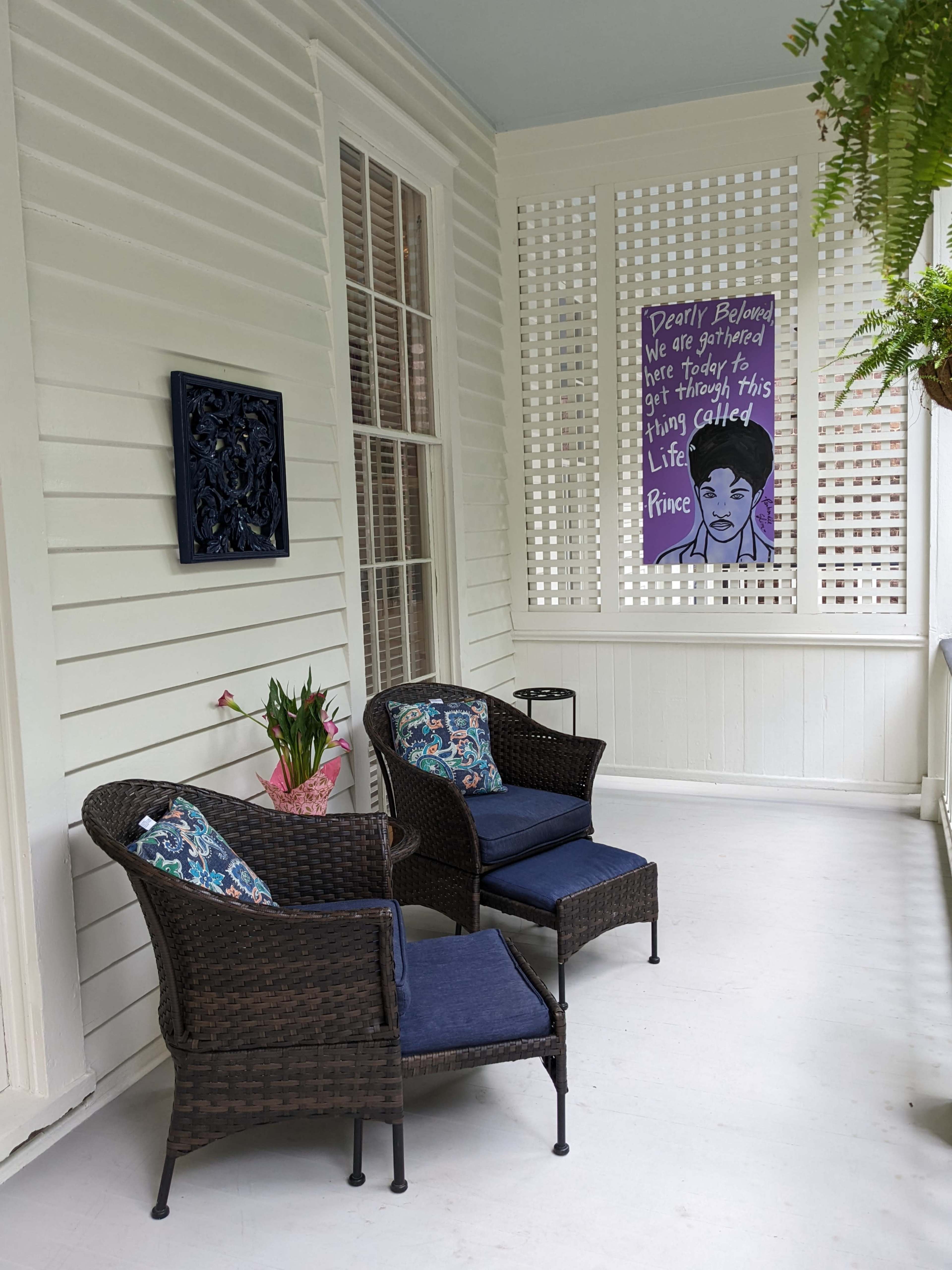 A porch features two wicker chairs with blue cushions, a plant, and a colorful artwork hanging on the wall.