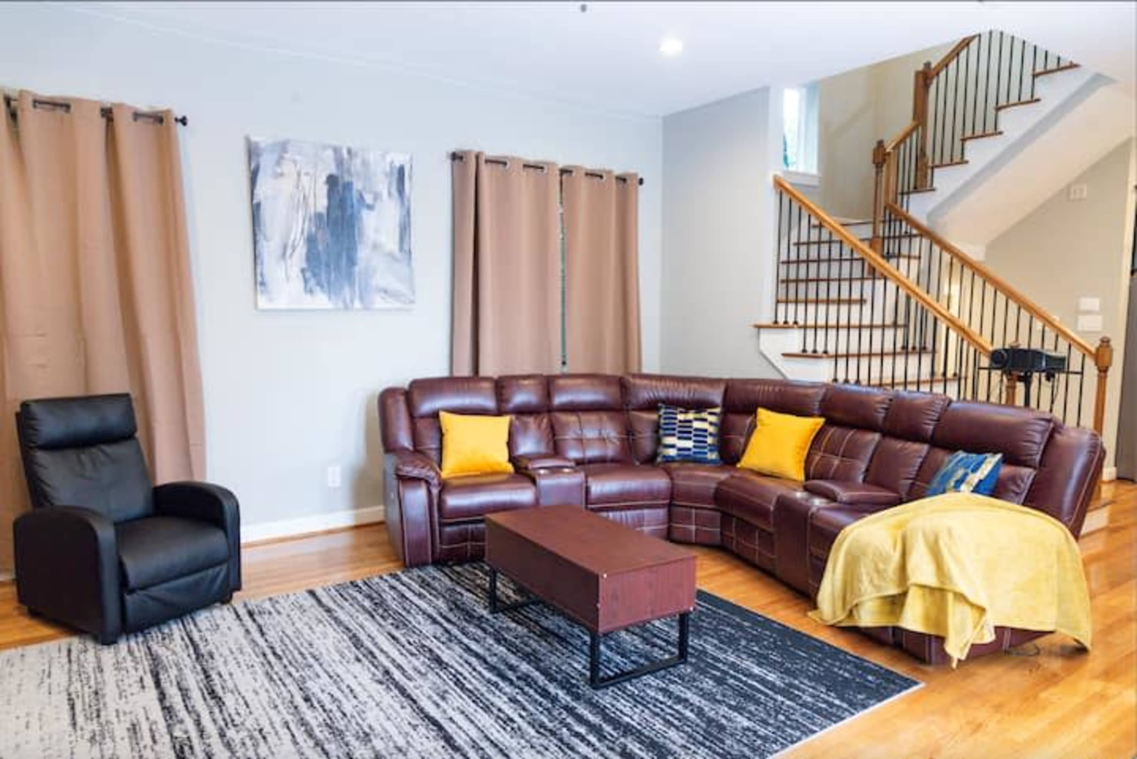 The image shows a living room with a brown sectional sofa, a black armchair, a wooden coffee table, a yellow throw blanket, and large curtains, along with wooden stairs leading to another level.