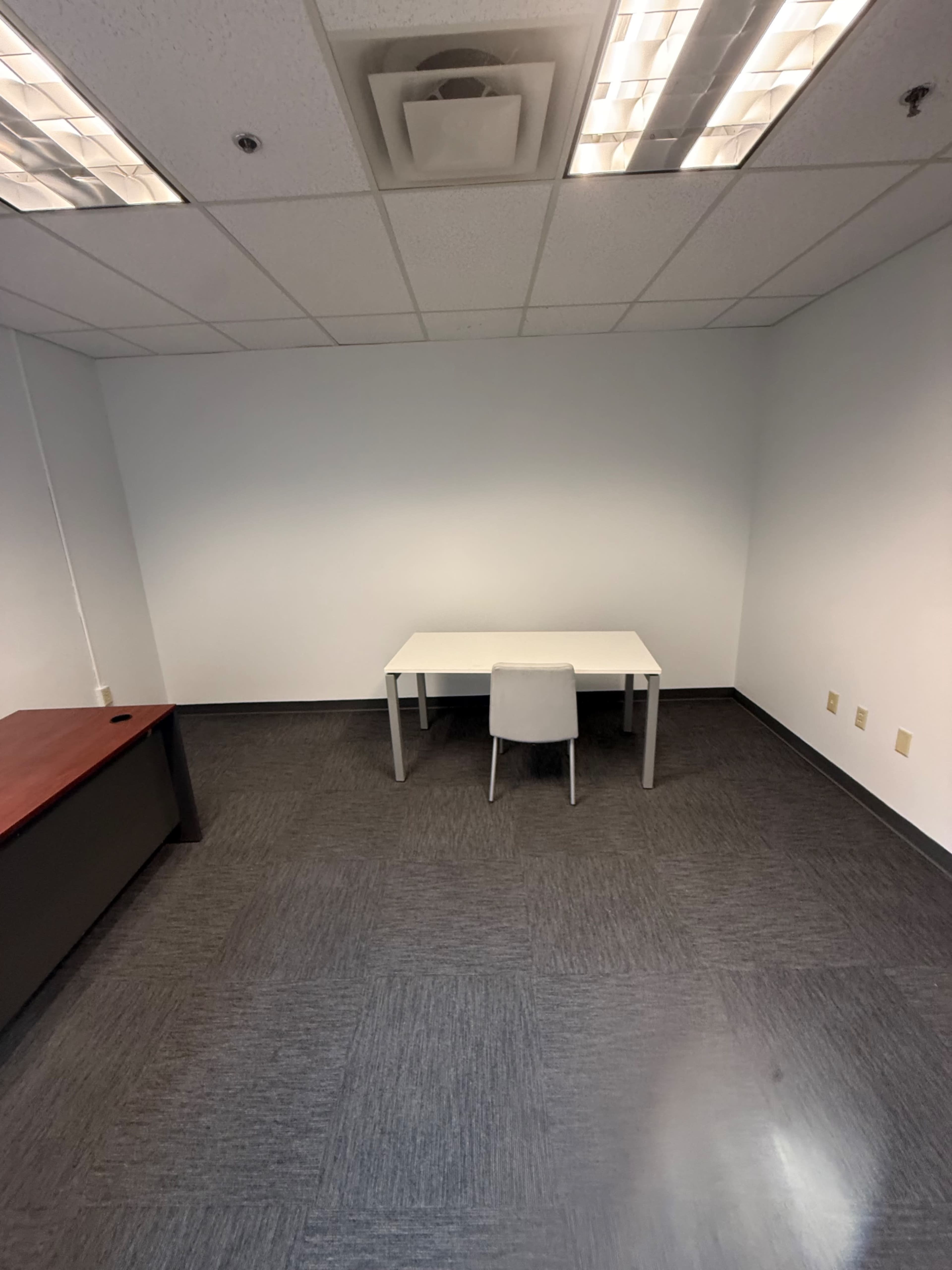 The image shows a sparse office space with a single white table and a chair, set against plain white walls and gray carpeting.