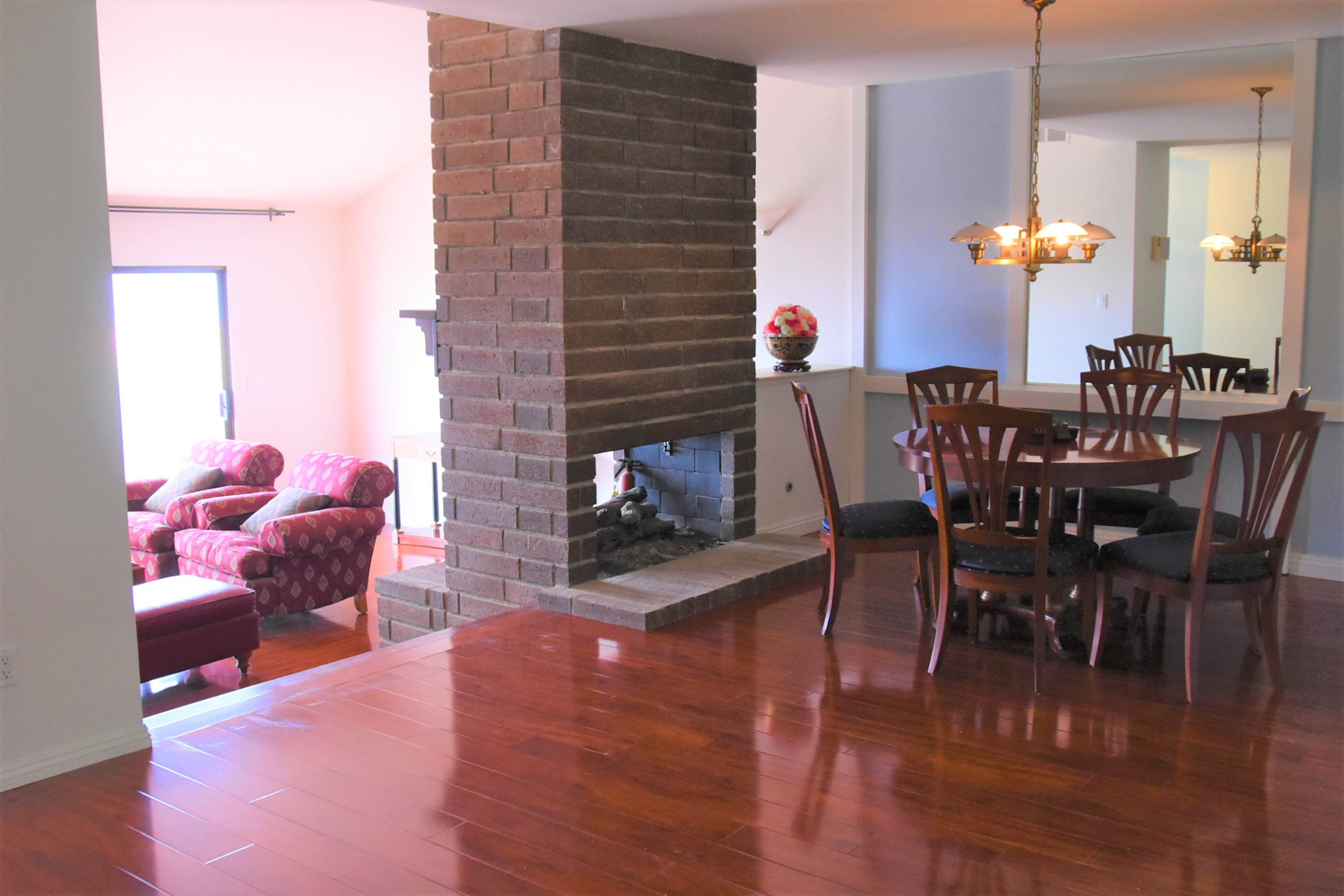 The image shows a living and dining area featuring a brick fireplace, wooden flooring, and a round dining table with chairs.