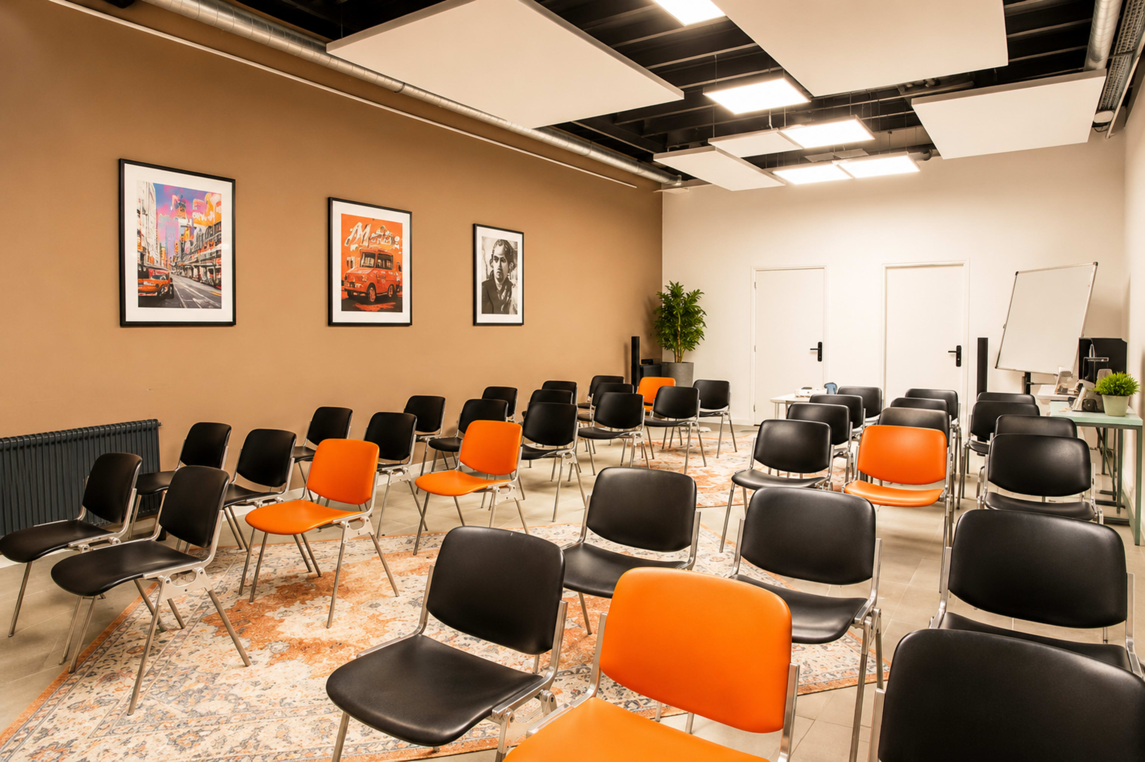 The image shows a sparsely furnished meeting room with rows of black and orange chairs arranged around a decorative rug, flanked by framed wall art and a whiteboard.
