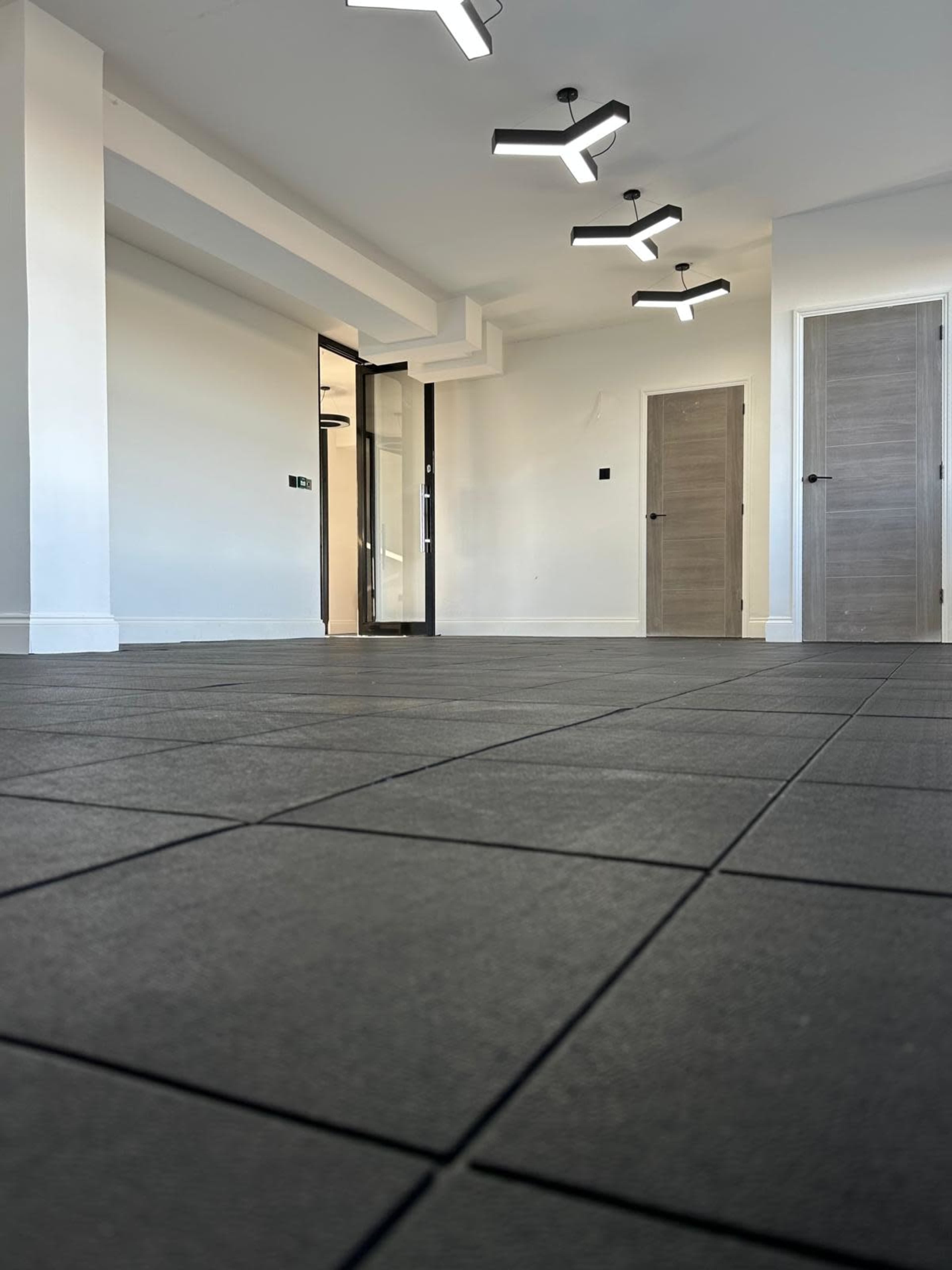 The image shows an empty room with black tiled flooring, featuring modern lighting fixtures and two closed doors leading to adjacent spaces.