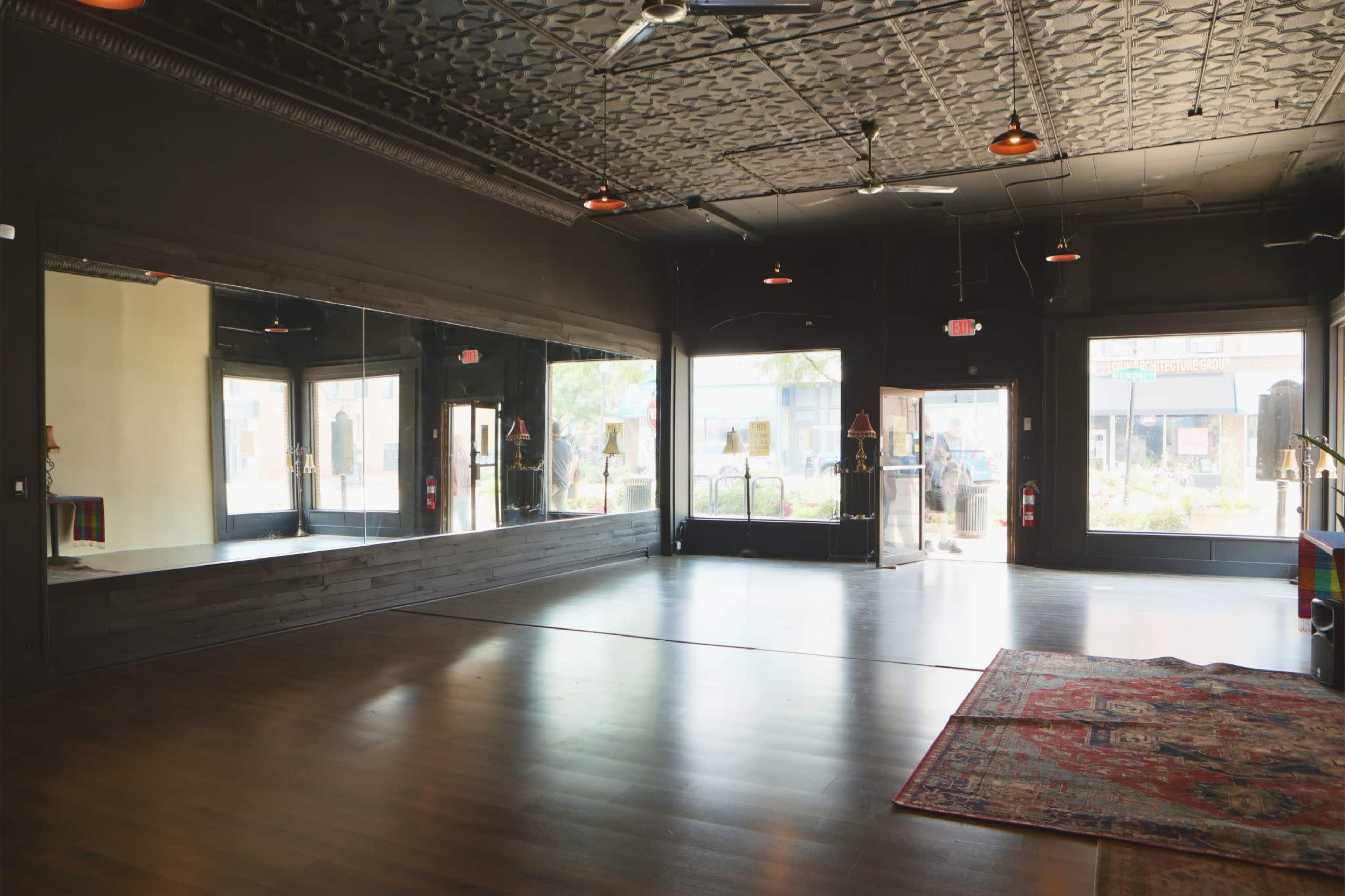 The image shows a spacious interior with dark wooden flooring, large windows, and a mirrored wall on one side.