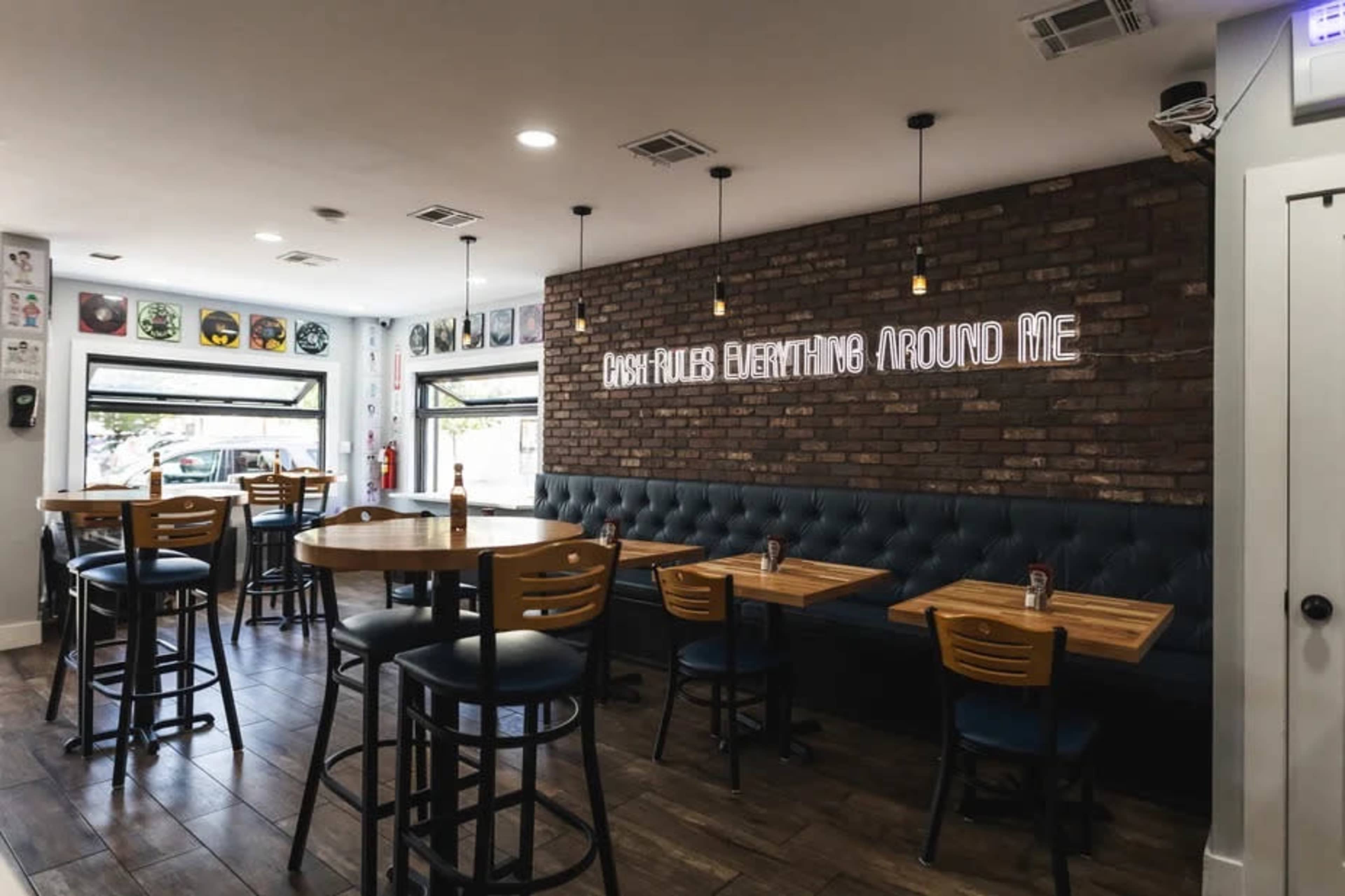 The image shows a modern restaurant interior with wooden tables and chairs, featuring a brick wall with neon signage and decorative artwork on the surrounding walls.