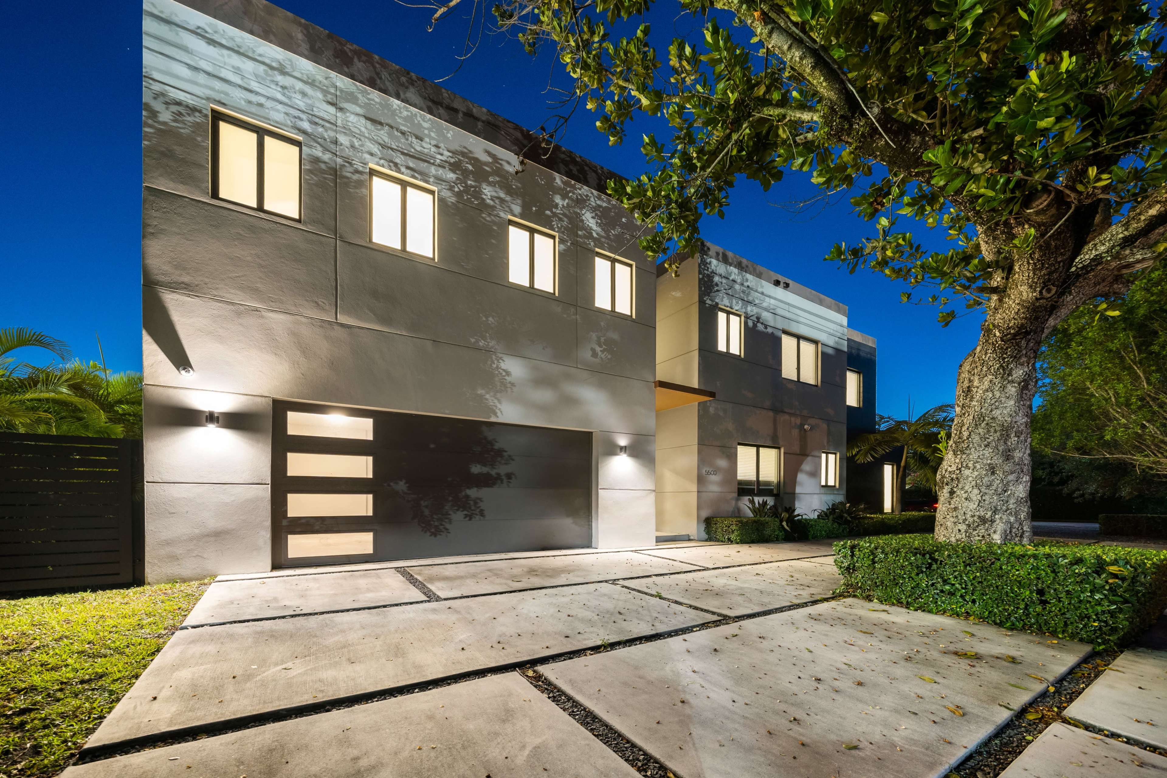 A modern two-story house with a garage and large windows is illuminated at dusk, surrounded by greenery.